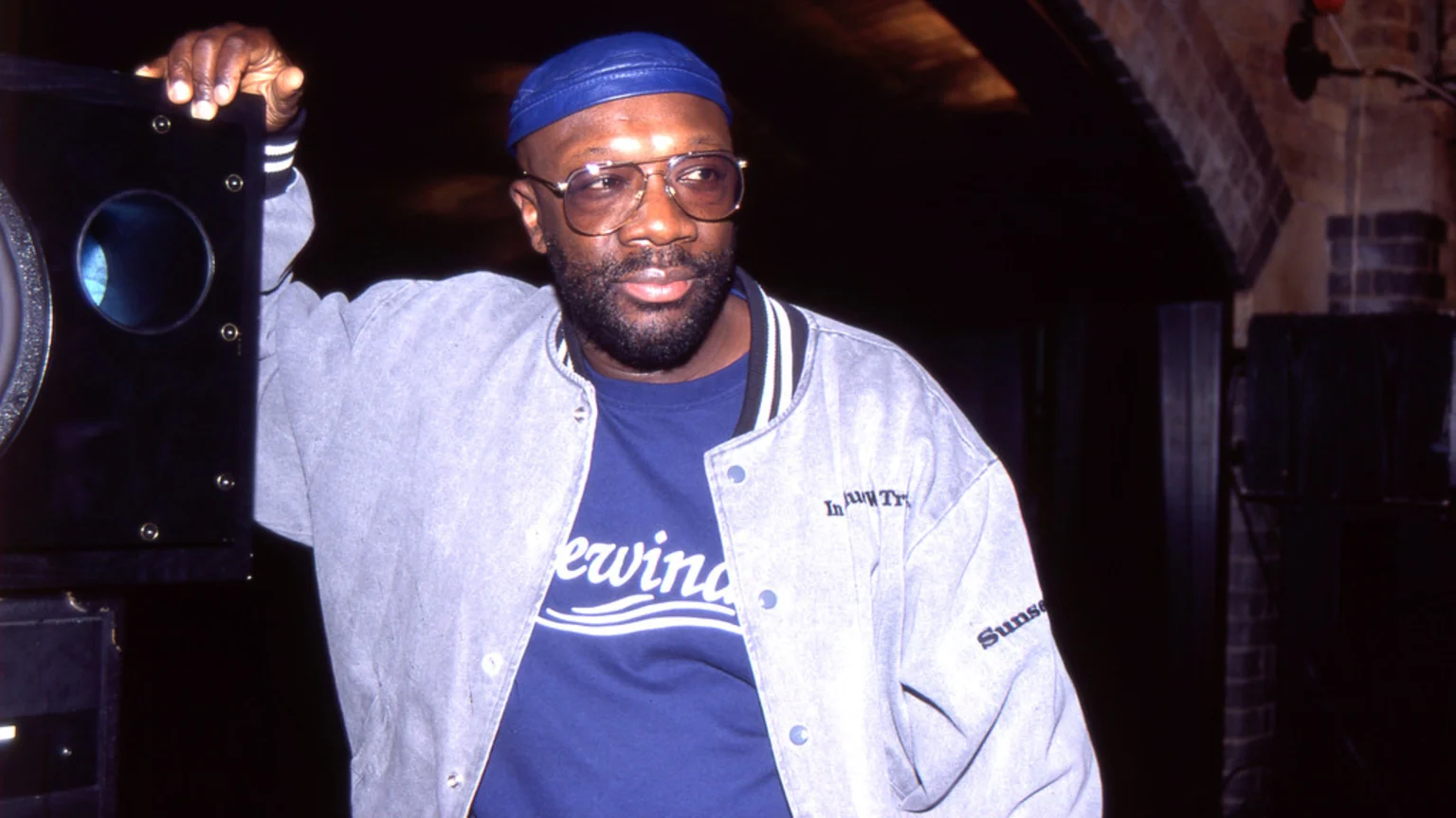 A dark-skinned man with a beard, blue cap, and glasses, wearing a grey jacket over a blue t-shirt, stands and looks to the side, with his hand on a large black speaker.
