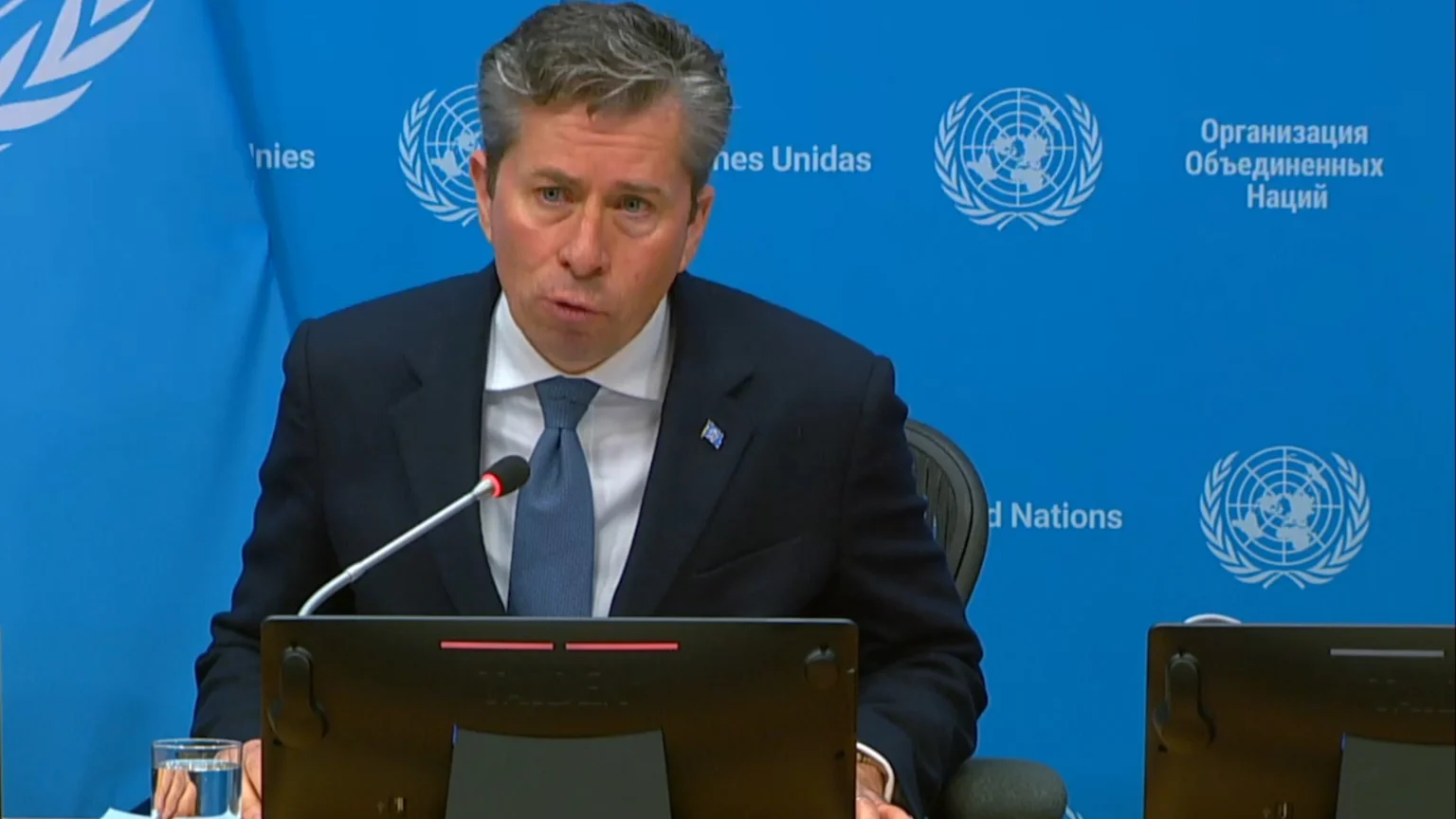 A man in a dark suit and blue tie speaks into a microphone at a desk, with a blue background featuring UN logos and text in multiple languages.