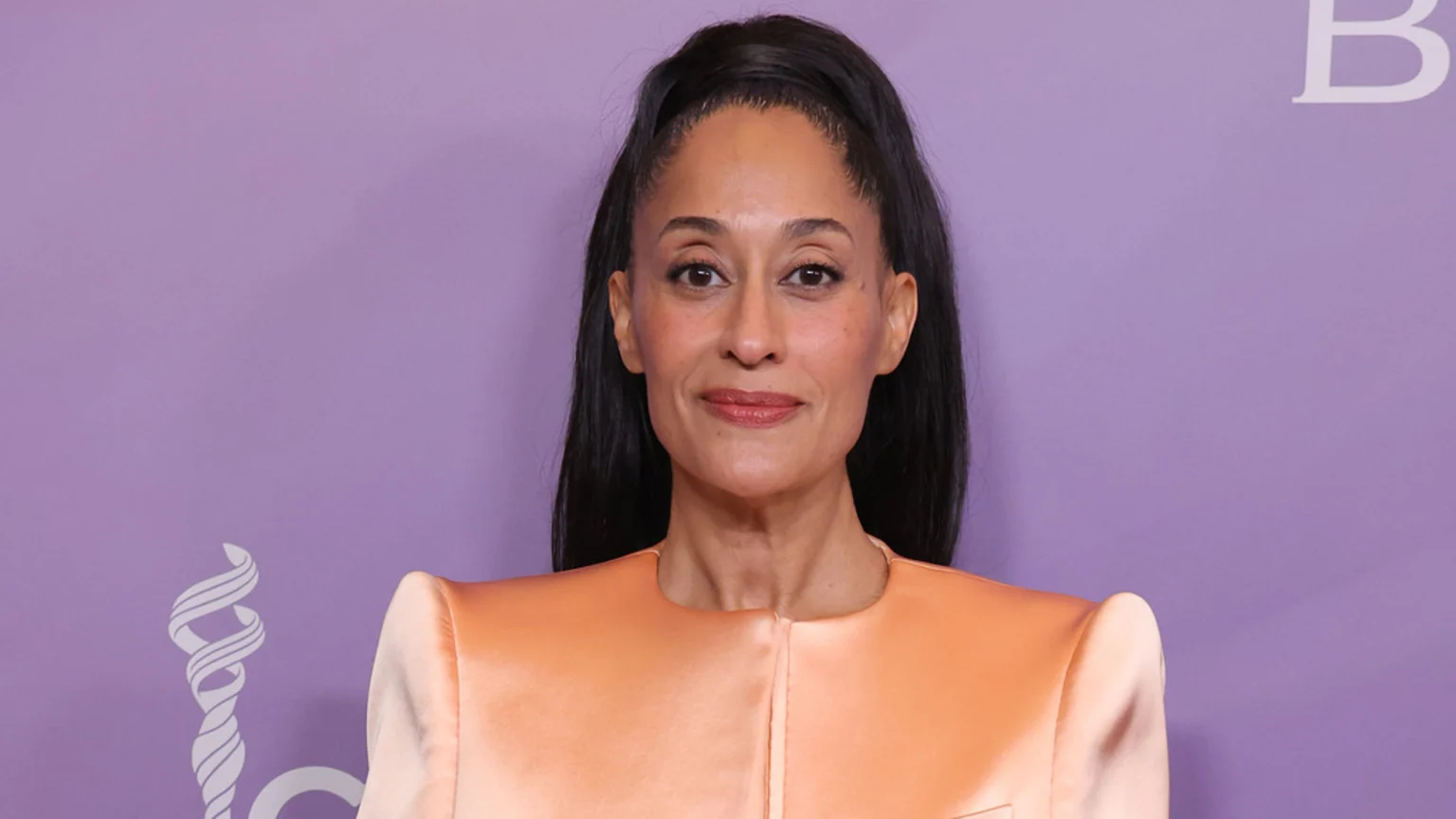 A woman with dark hair styled in a high ponytail, wearing a peach-orange blazer, gazes forward with a faint smile against a lavender background.