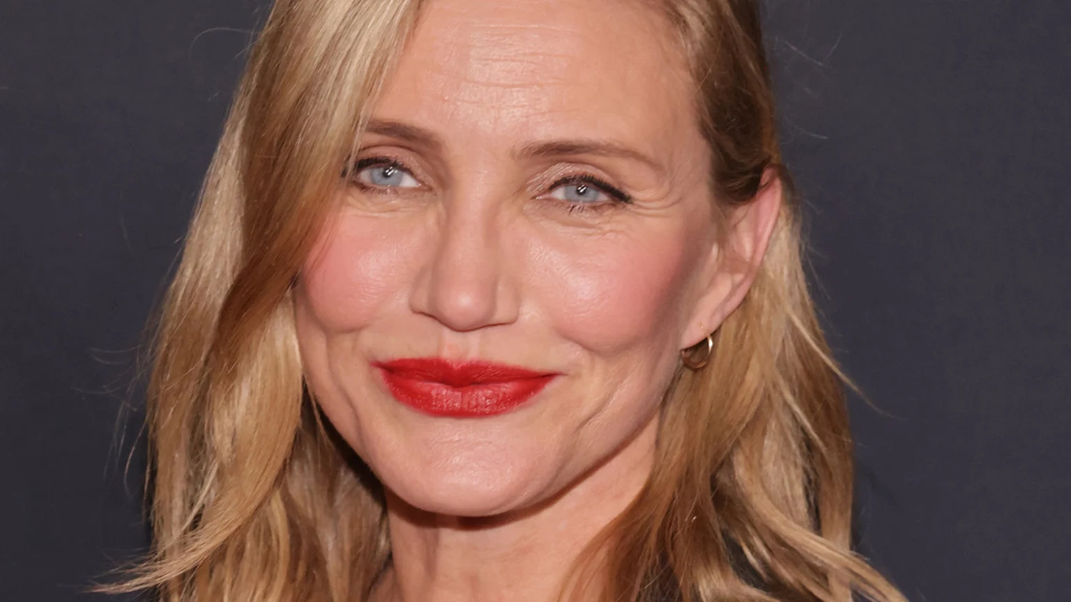 A close-up portrait of Cameron Diaz with blonde hair, blue eyes, red lipstick, and a gentle smile, showing visible facial lines against a dark background.