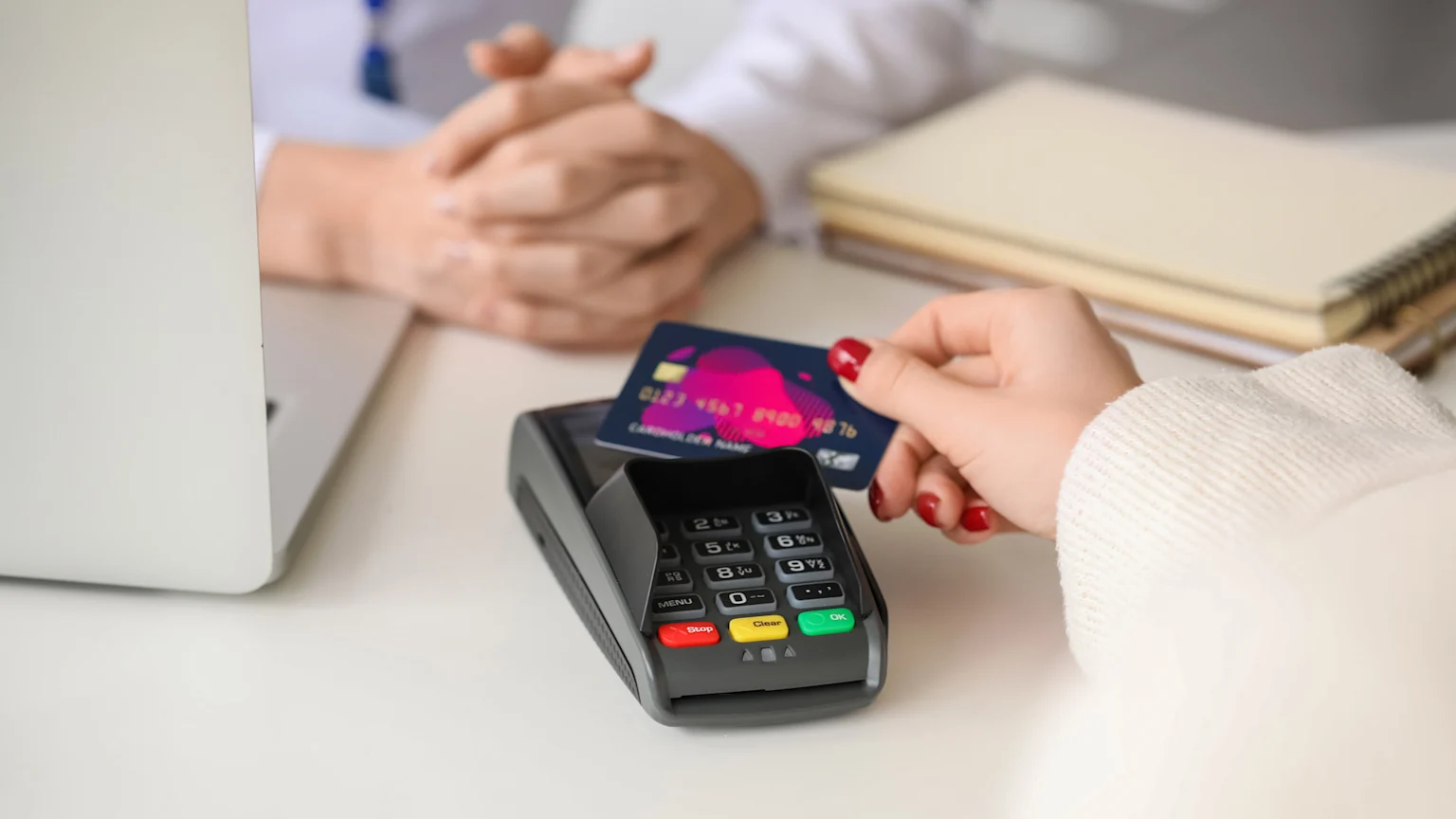 A person holds a dark blue credit card over a gray payment terminal on a white desk, with a laptop and a second person's hands blurred in the background.