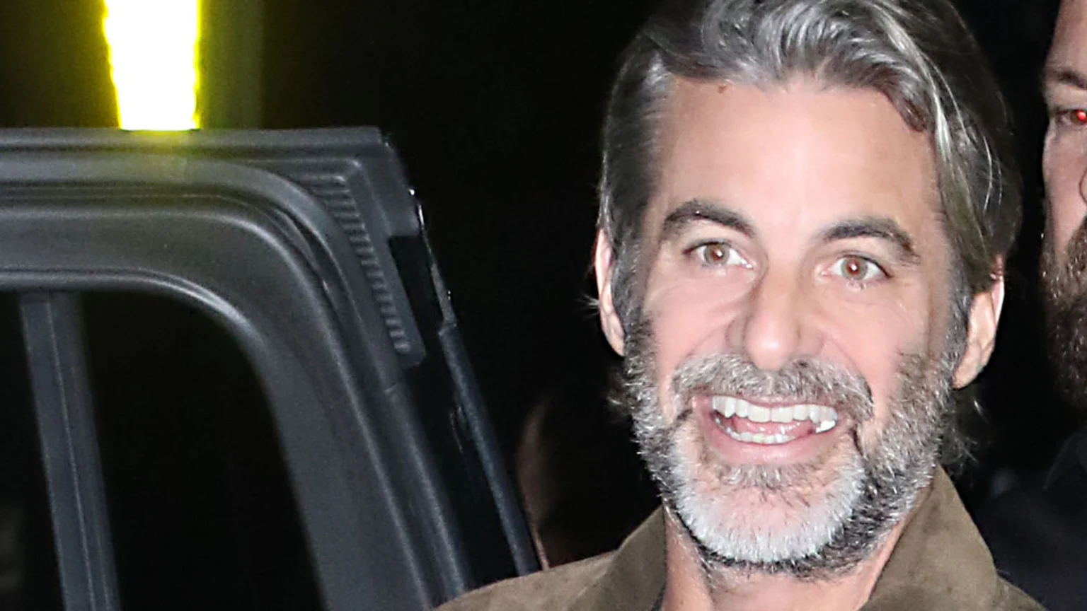 A man with graying hair and beard, hazel eyes, and a wide smile wears a brown jacket, with a partial face of another person and a car door visible in the dark background.