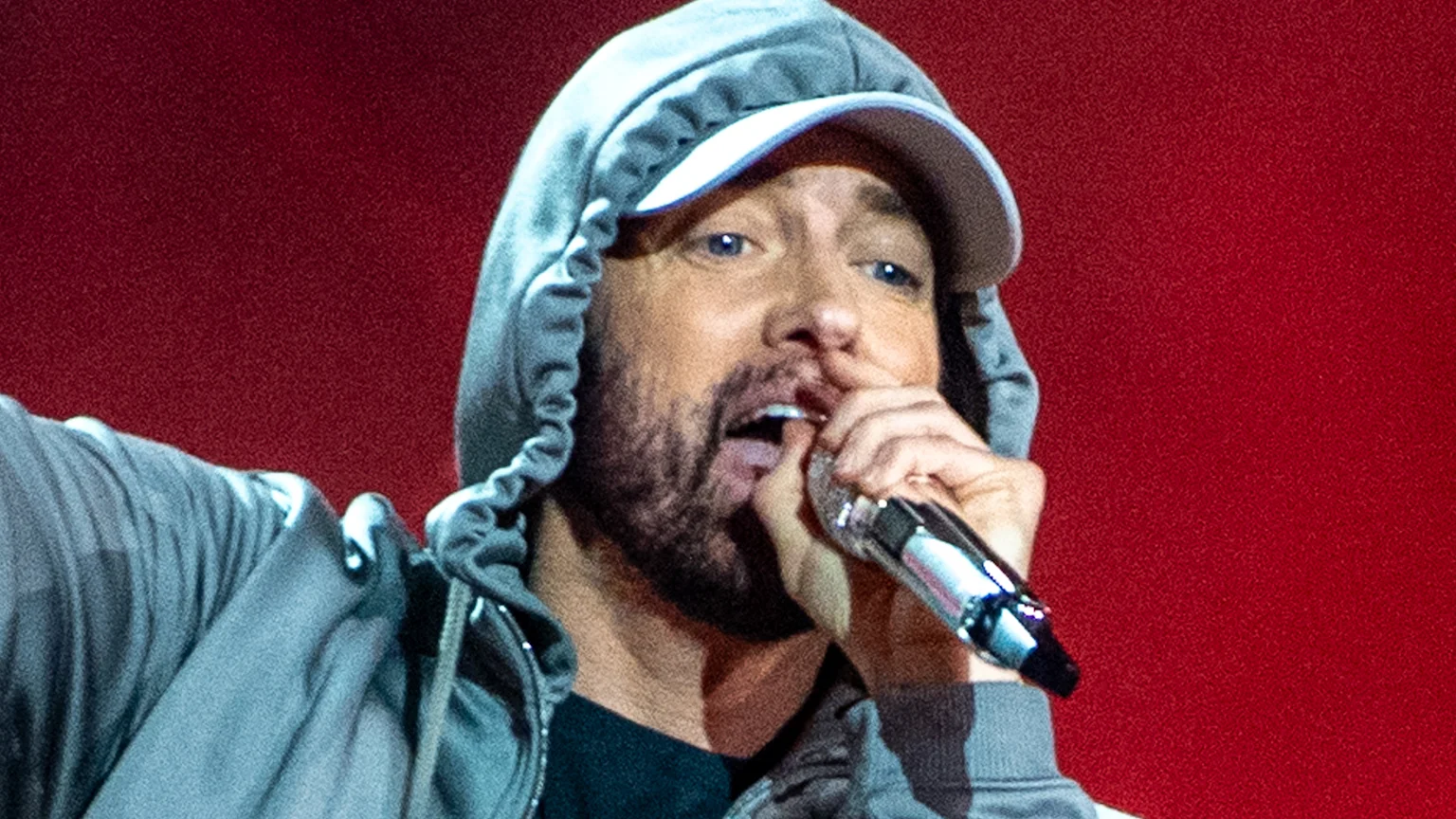 Eminem in an olive Kangol cap, white t-shirt, and silver chain, looks slightly left with an in-ear monitor, against a black background.