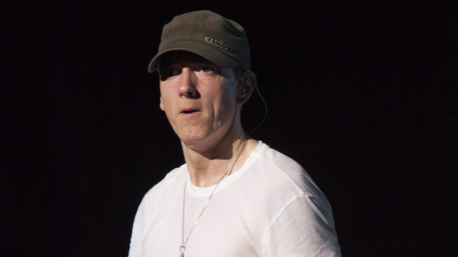 Eminem in an olive Kangol cap, white t-shirt, and silver chain, looks slightly left with an in-ear monitor, against a black background.