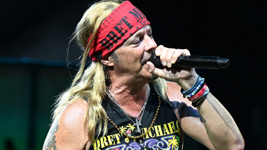 Bret Michaels, with long blonde hair and a red bandana, sings into a microphone while wearing a black tank top and multiple necklaces and bracelets.