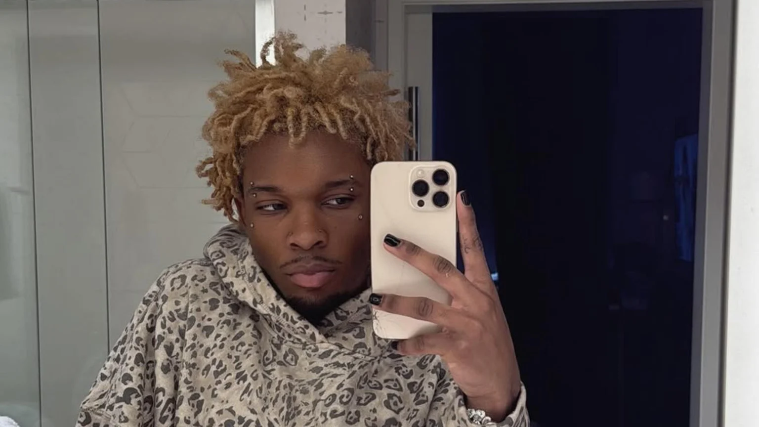Singer D4vd takes a mirror selfie, showing off blonde dreadlocks, facial piercings, black nail polish, and an animal print hoodie.