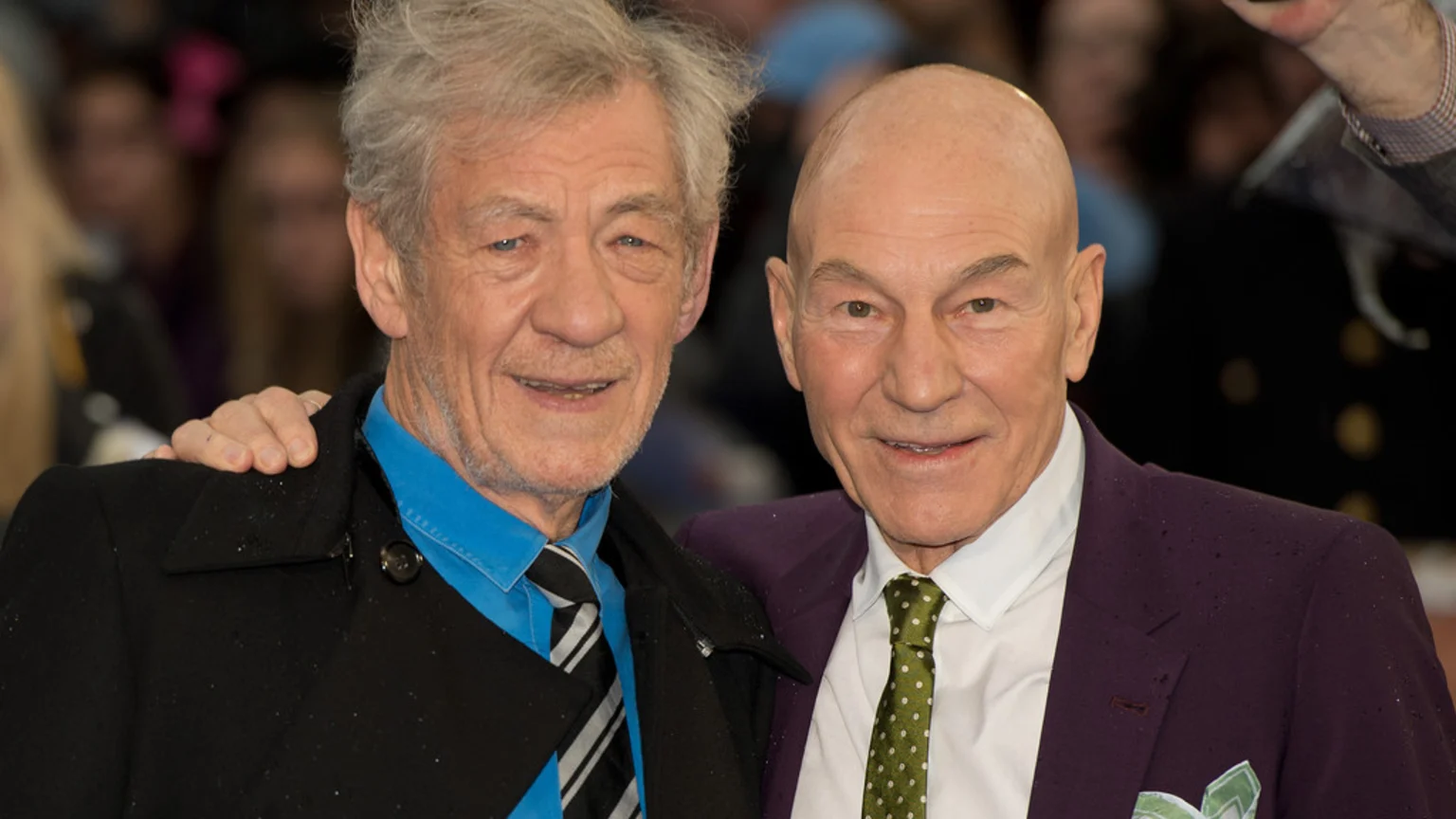 Actors Ian McKellen (left), in a blue shirt, and Patrick Stewart (right), in a purple blazer, smile for the camera, with McKellen's hand on Stewart's shoulder.
