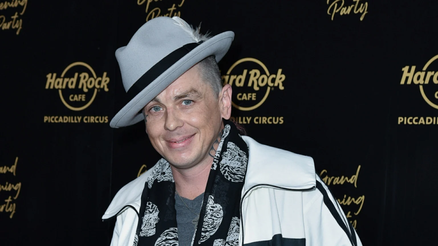 A man with facial tattoos and a short haircut, wearing a light grey fedora with a feather, a black and white patterned scarf, and a white jacket, smiles at the camera in front of a "Hard Rock Cafe Piccadilly Circus" backdrop.