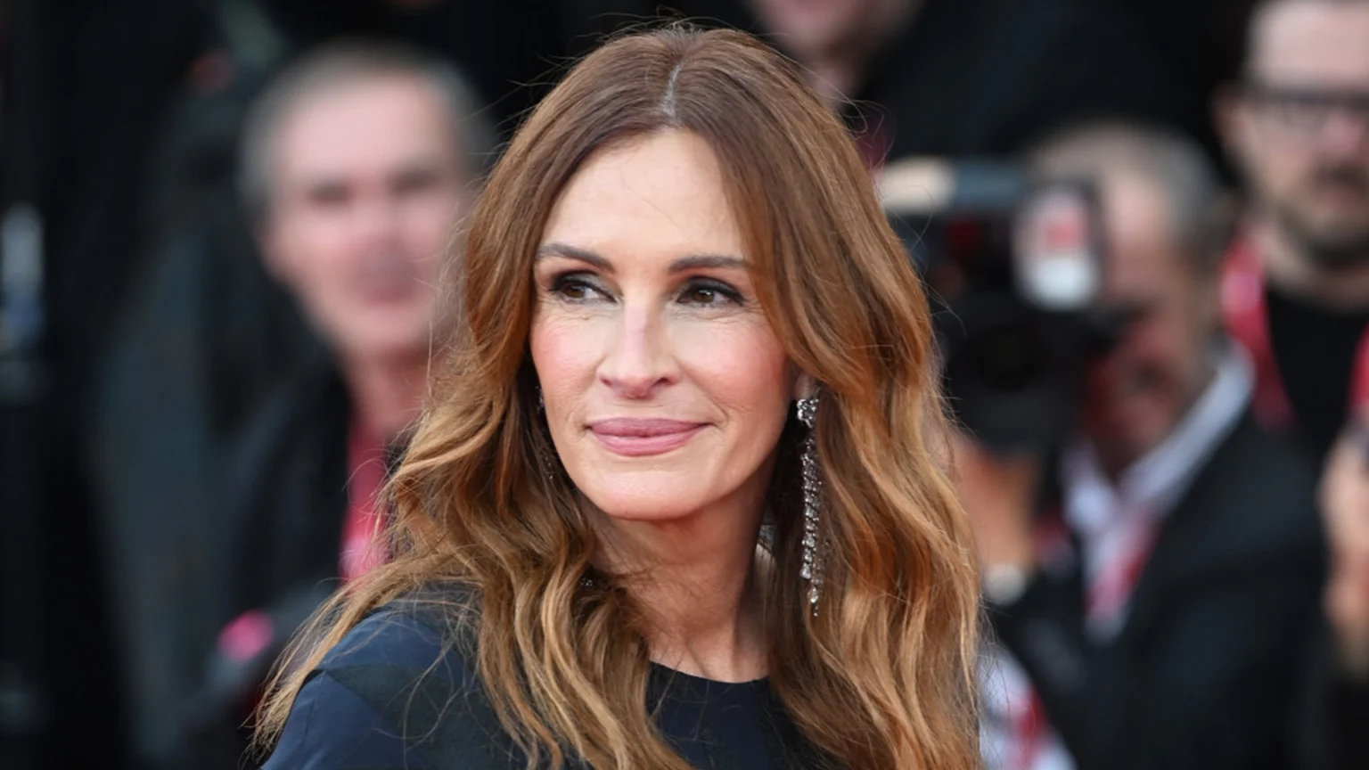 A close-up of actress Julia Roberts, with long wavy brown hair and sparkling earrings, looking to her right with a gentle smile against a blurred background of people.