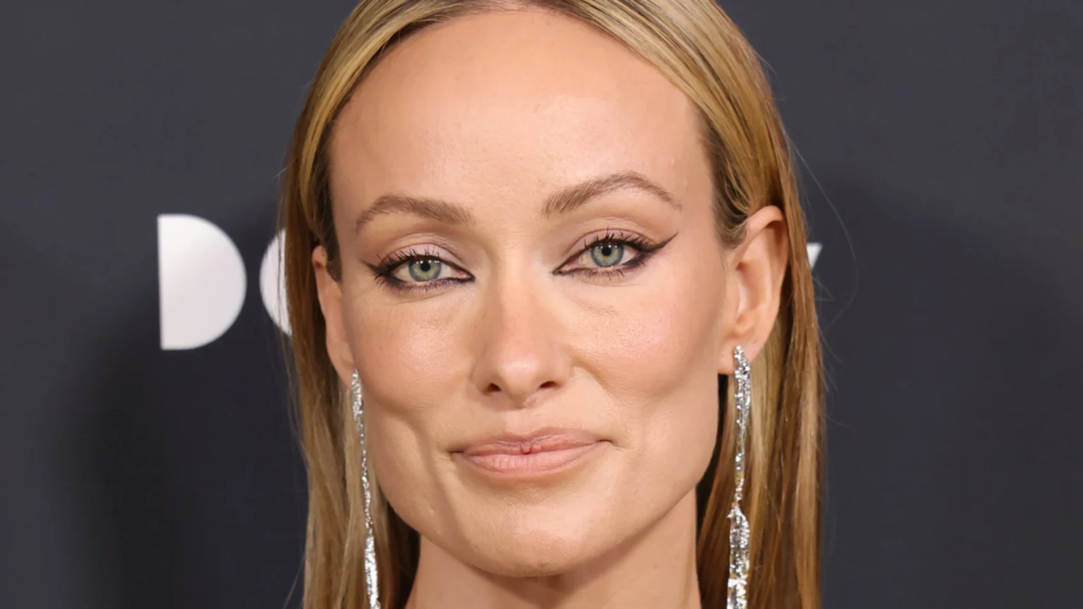 Close-up of Olivia Wilde with blonde hair, light eyes, and winged eyeliner, wearing long sparkly earrings, against a dark grey background with a partial white logo.