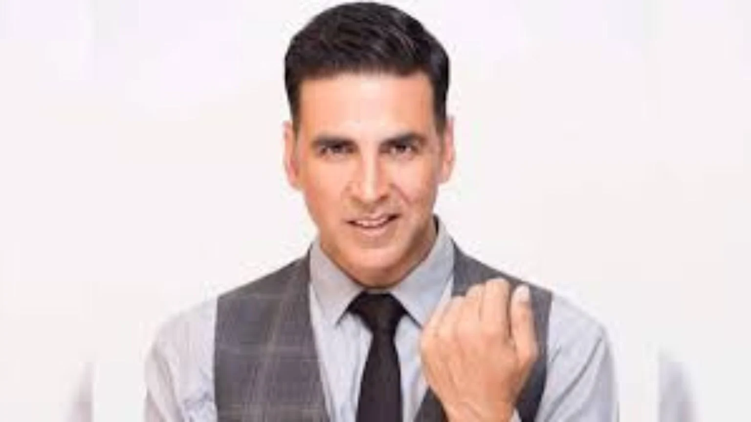Bollywood actor Akshay Kumar smiles, wearing a grey shirt, checkered vest, and black tie, looking at the camera with his right hand raised.