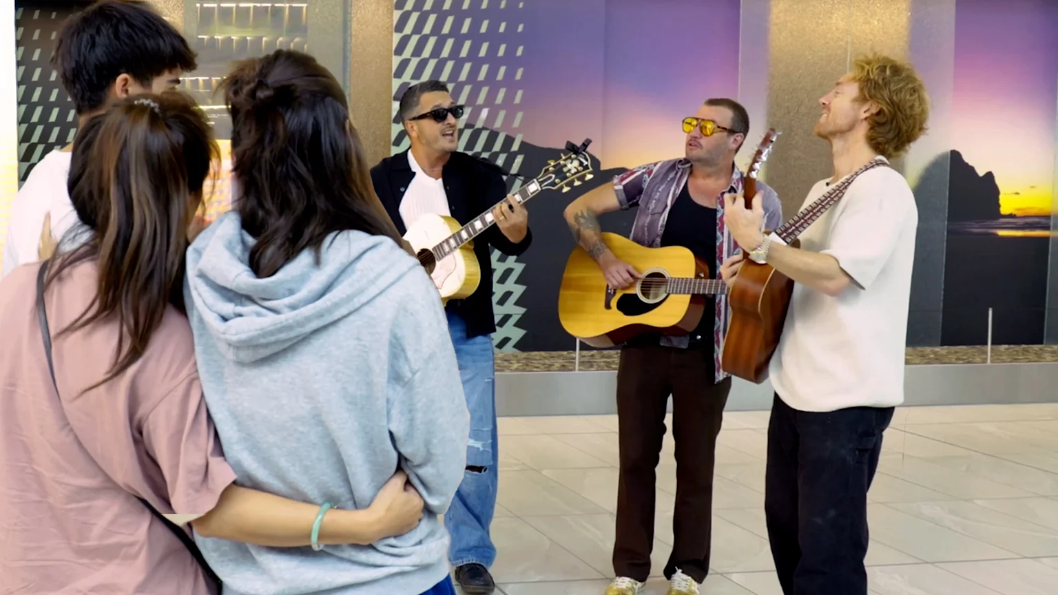 The image shows five adults indoors: three men playing acoustic guitars and singing, and two individuals in casual clothing observing. The backdrop features modern geometric wall art and a large mural of a sunset landscape.
