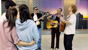 The image shows five adults indoors: three men playing acoustic guitars and singing, and two individuals in casual clothing observing. The backdrop features modern geometric wall art and a large mural of a sunset landscape.