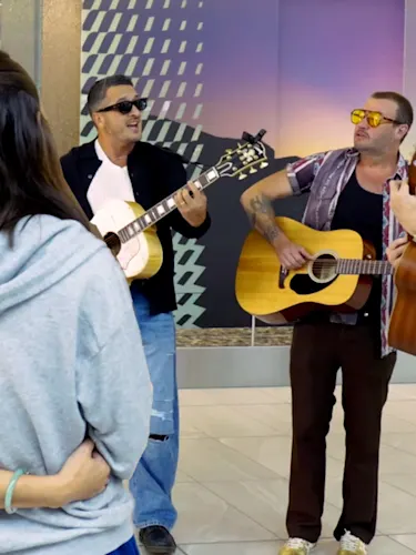 The image shows five adults indoors: three men playing acoustic guitars and singing, and two individuals in casual clothing observing. The backdrop features modern geometric wall art and a large mural of a sunset landscape.