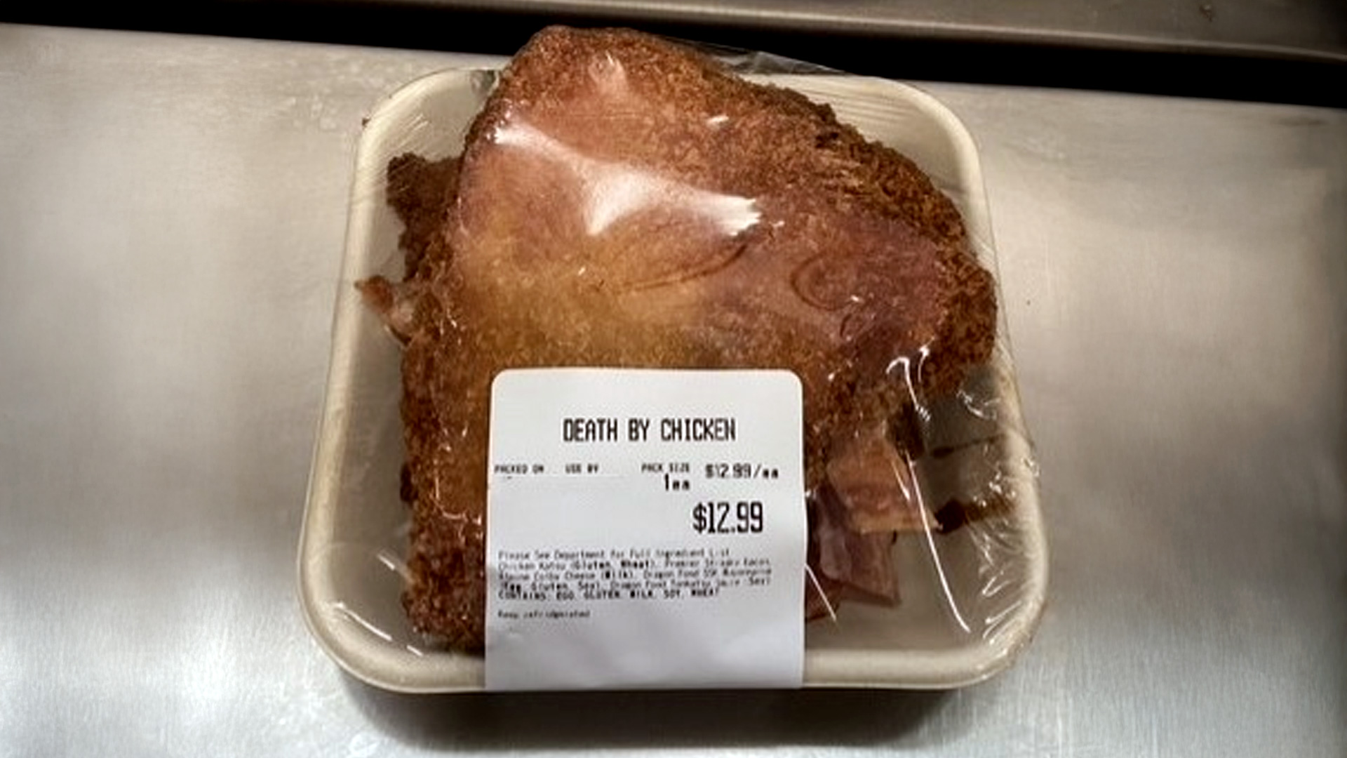 A packaged chicken katsu dish labeled "DEATH BY CHICKEN" in a plastic-wrapped foam tray, priced at $12.99.