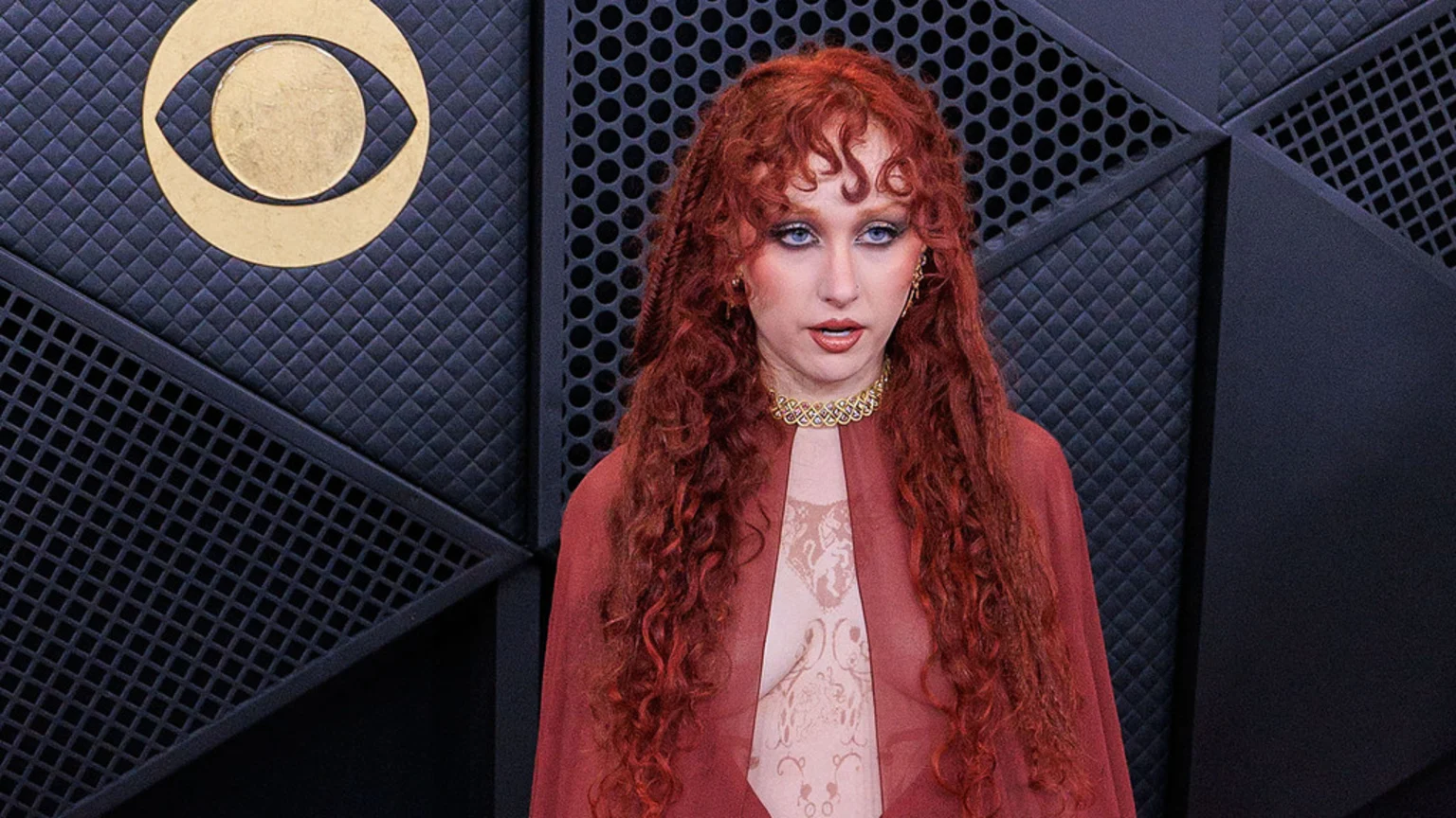 A close-up portrait of a person with long curly red hair, dramatic blue eyeshadow and red lipstick, wearing a red cape-like garment, sheer patterned top, and gold jewelry, standing in front of a dark geometric textured wall with a gold CBS Eye logo. The person looks directly at the viewer with a neutral expression in an indoor event space.