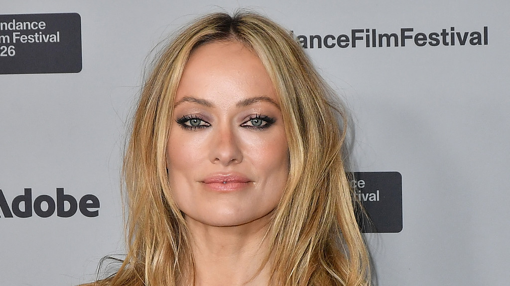 A close-up of actress Olivia Wilde with long blonde hair, smokey eye makeup, and a small labret piercing, looking forward against a light grey background with "Sundance Film Festival" and "Adobe" logos.