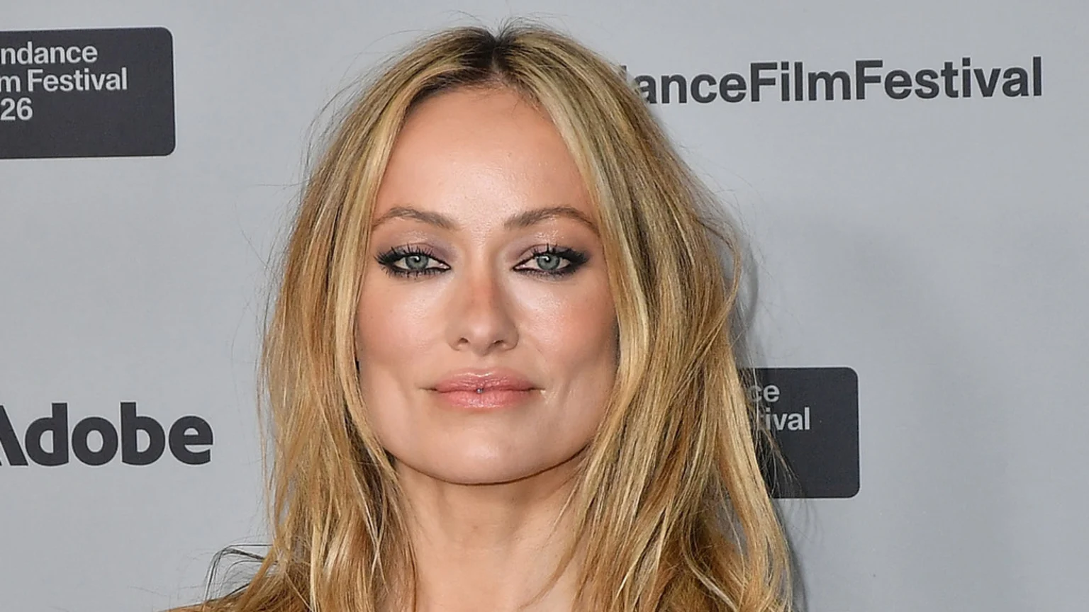 A close-up of actress Olivia Wilde with long blonde hair, smokey eye makeup, and a small labret piercing, looking forward against a light grey background with "Sundance Film Festival" and "Adobe" logos.