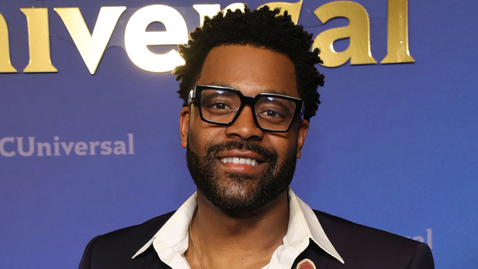 LaRoyce Hawkins smiles at the camera, wearing black glasses, a dark jacket, and a white shirt, against a blue background with "Universal" in gold and "CUniversal" in white.