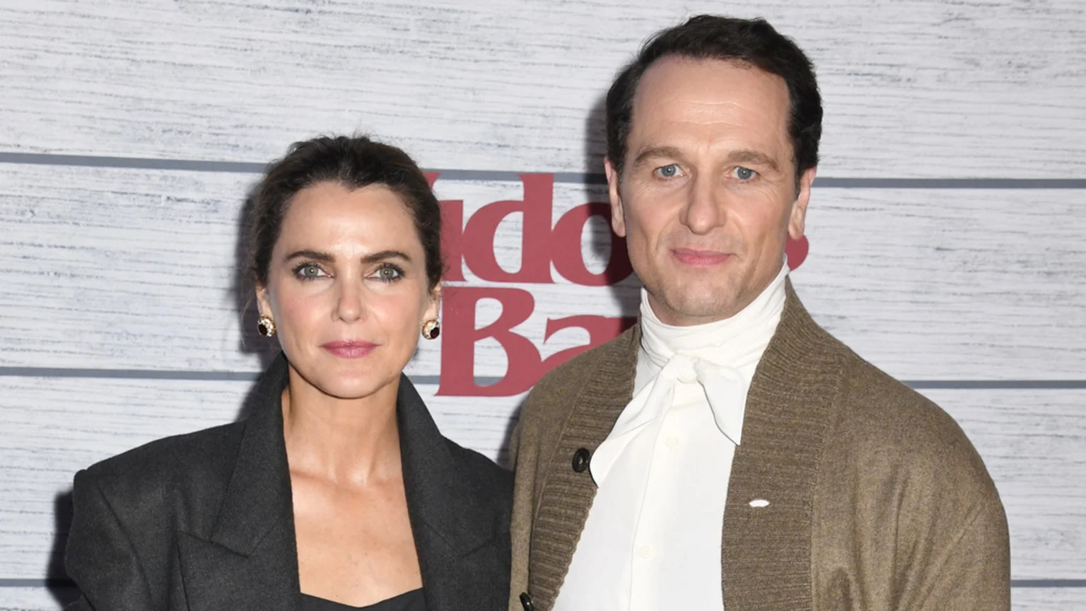 Keri Russell and Matthew Rhys stand side-by-side, both looking at the camera, Keri in a dark jacket and Matthew in a white shirt and brown cardigan, against a white wooden wall with red text.