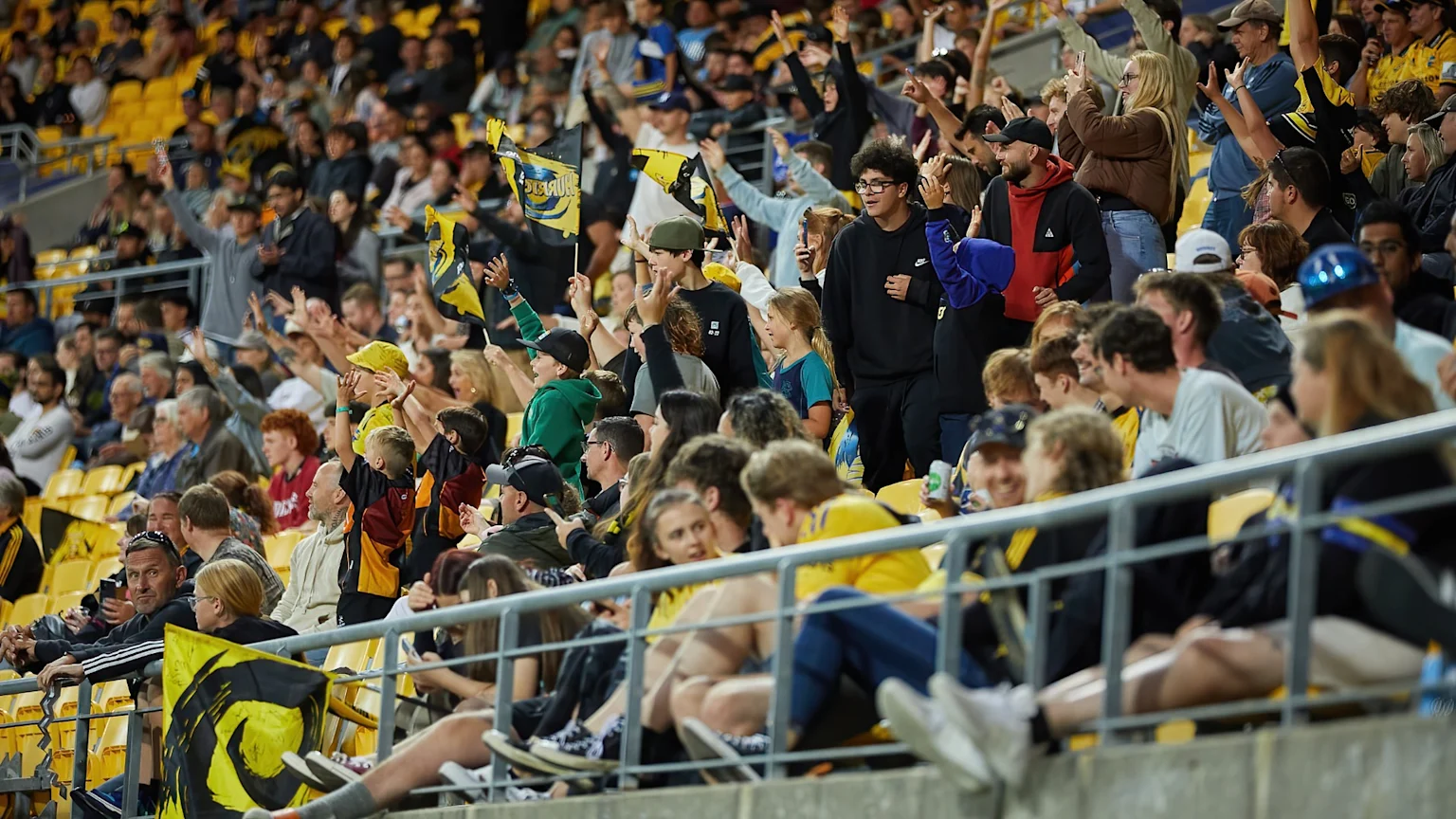 A large crowd of people in a stadium, many wearing yellow and black, cheer and raise their hands and flags, filling the yellow seats.