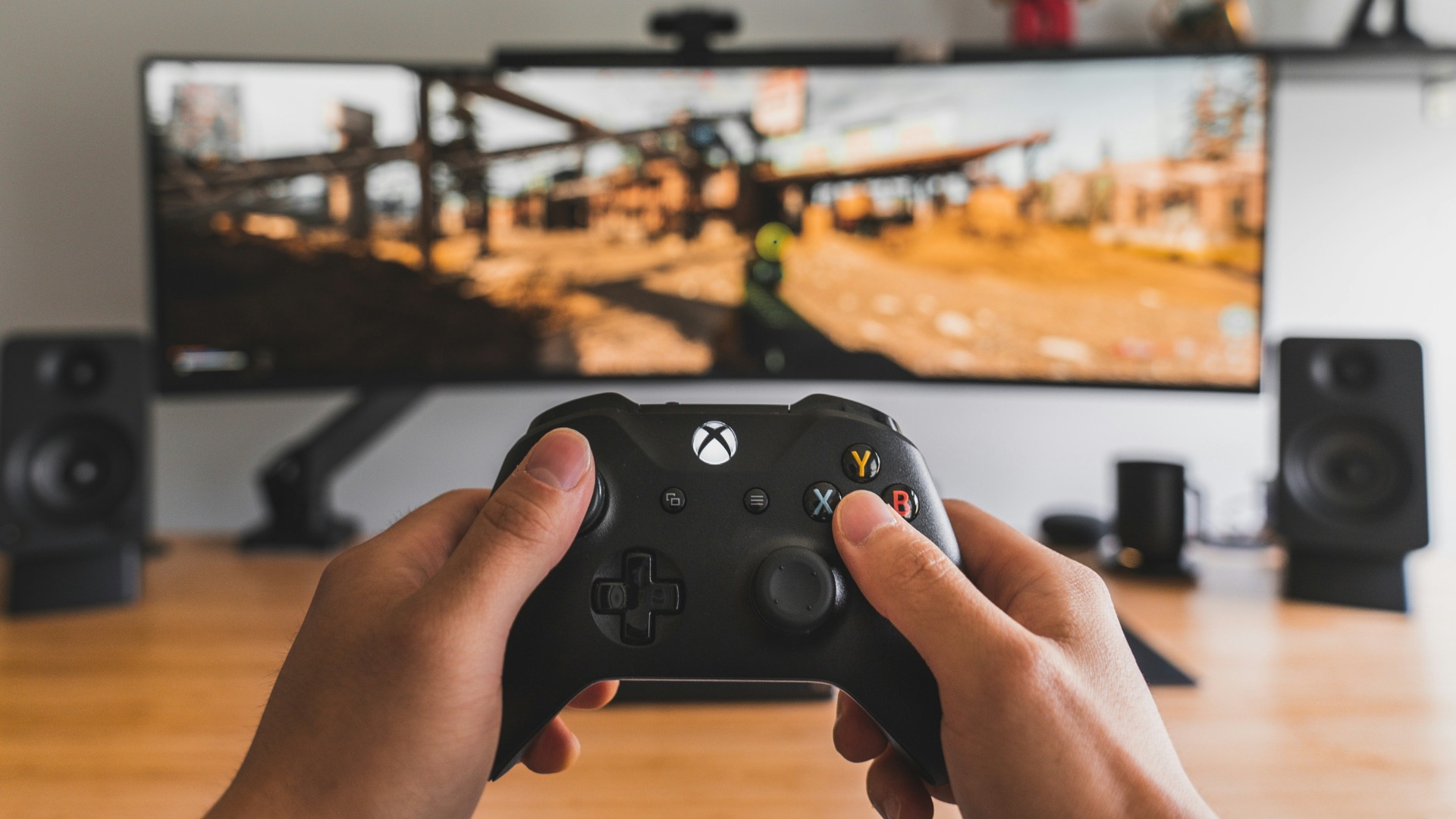 A person's hands holding a black Xbox controller are in the foreground, with a video game displayed on a large curved monitor and two speakers visible in the blurred background.