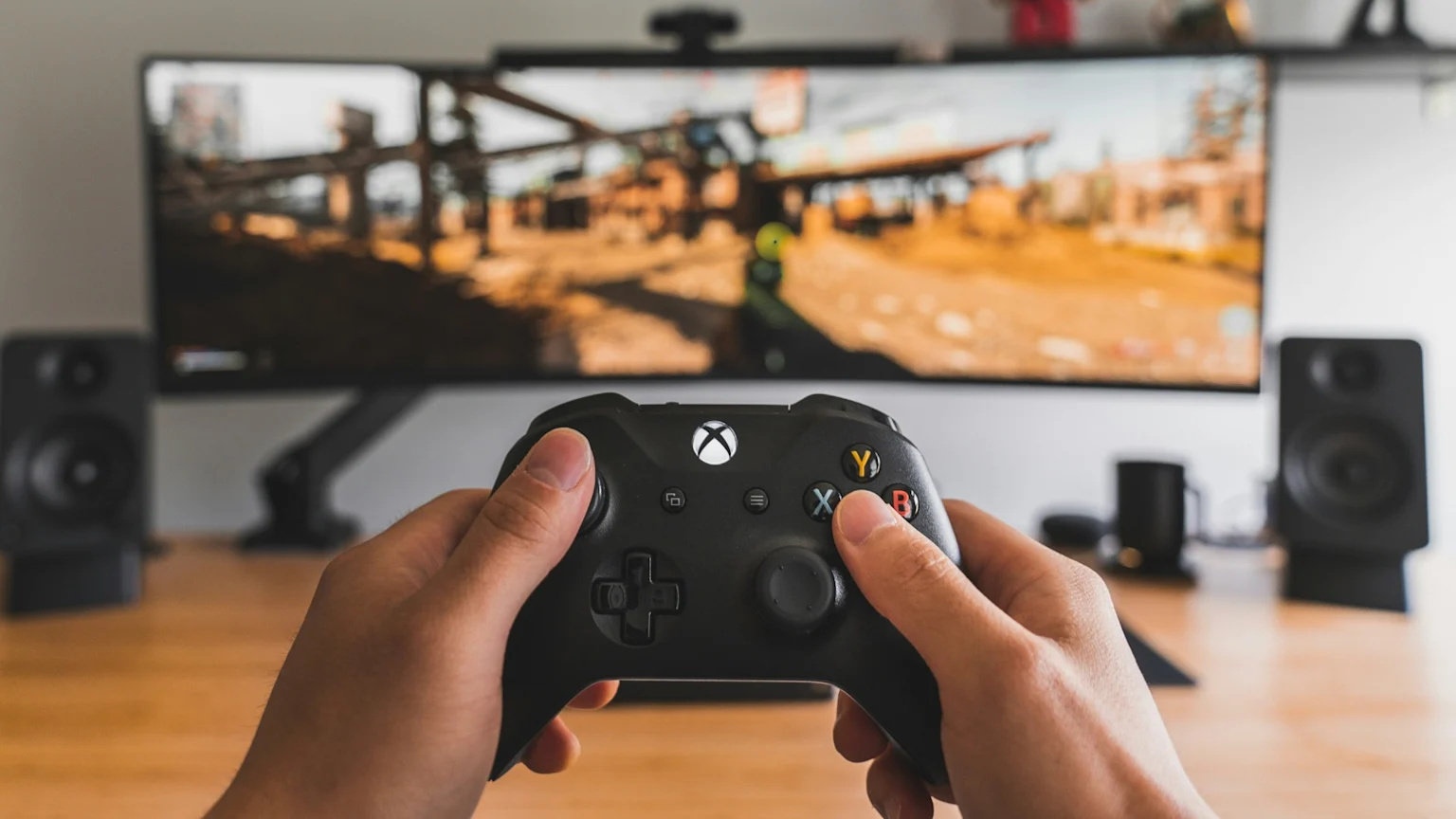 A person's hands holding a black Xbox controller are in the foreground, with a video game displayed on a large curved monitor and two speakers visible in the blurred background.