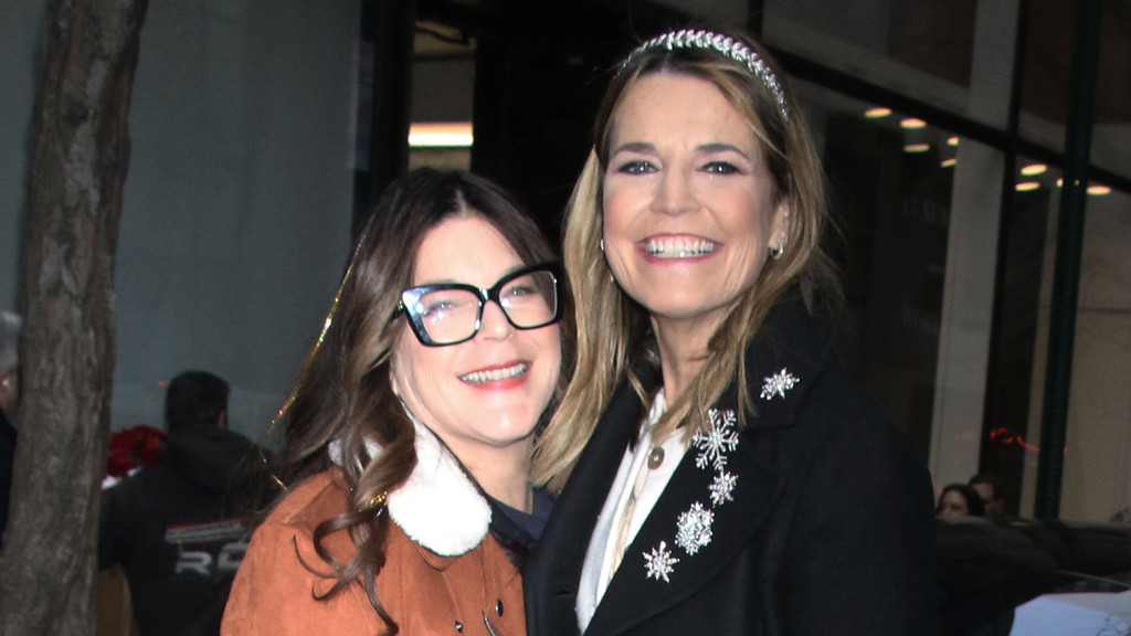 Two smiling women posing closely. The woman on the left has dark hair, wears large black glasses, and a brown, fur-collared jacket. The woman on the right has blonde hair, wears a sparkly headband, and a black coat embellished with silver snowflakes.