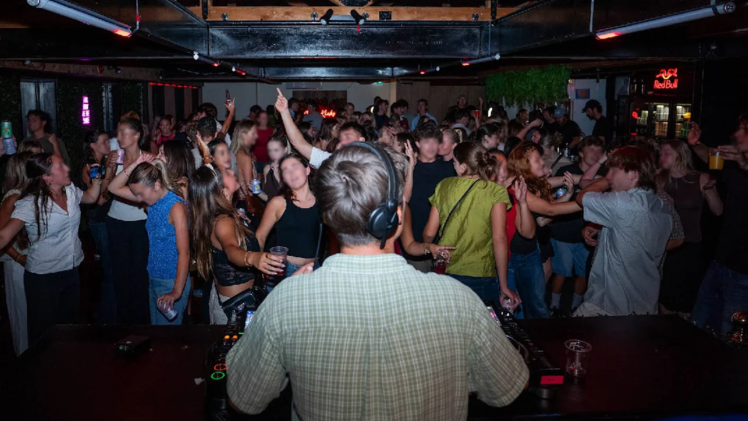 A DJ with headphones on stands with their back to the camera, facing a lively crowd dancing in a dimly lit music venue, with many people holding drinks.