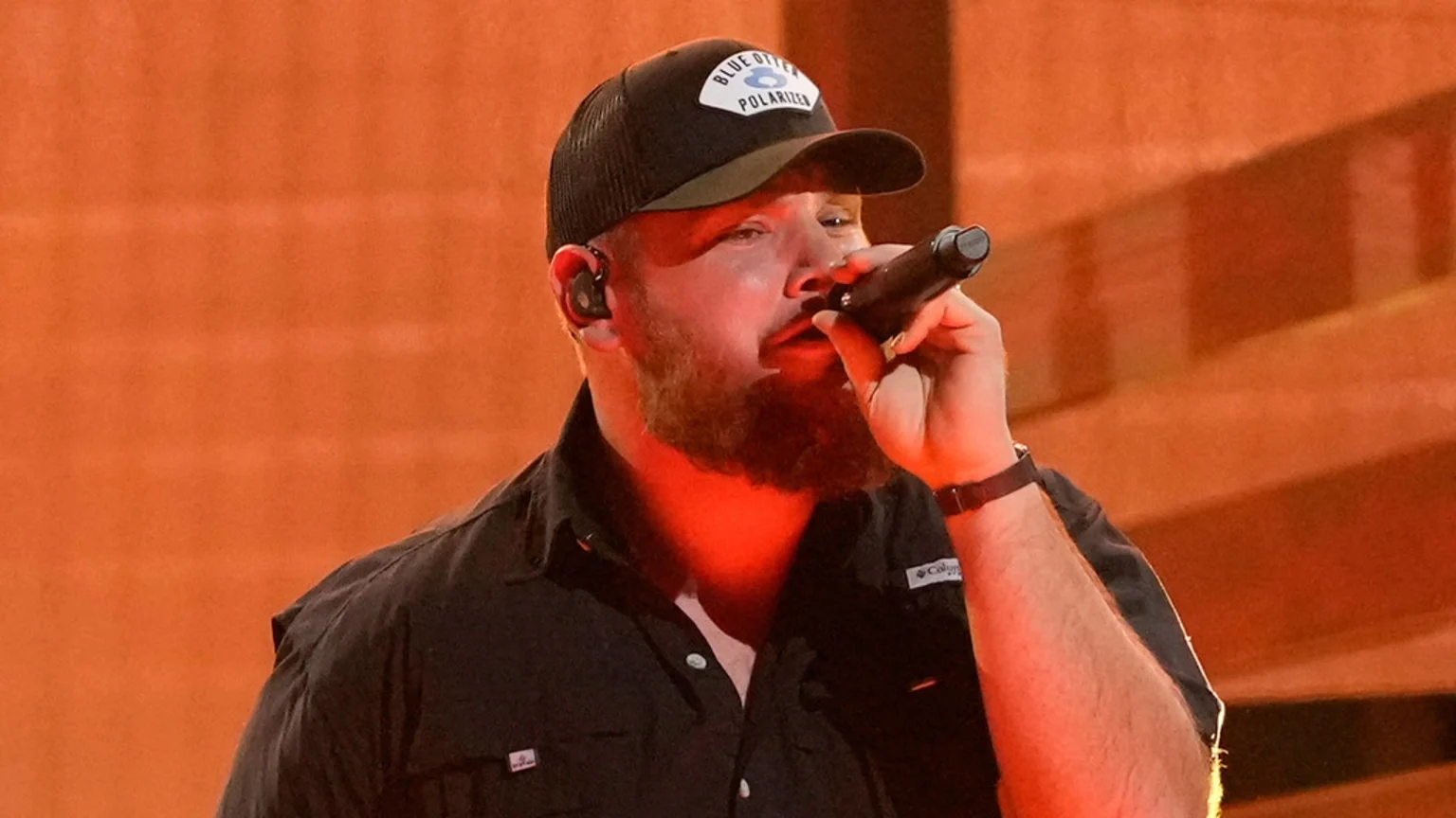 Luke Combs, a bearded man in a dark Columbia shirt and a baseball cap with a "Blue Otter Polarizer" logo, sings into a microphone, illuminated by warm orange light against an orange background.