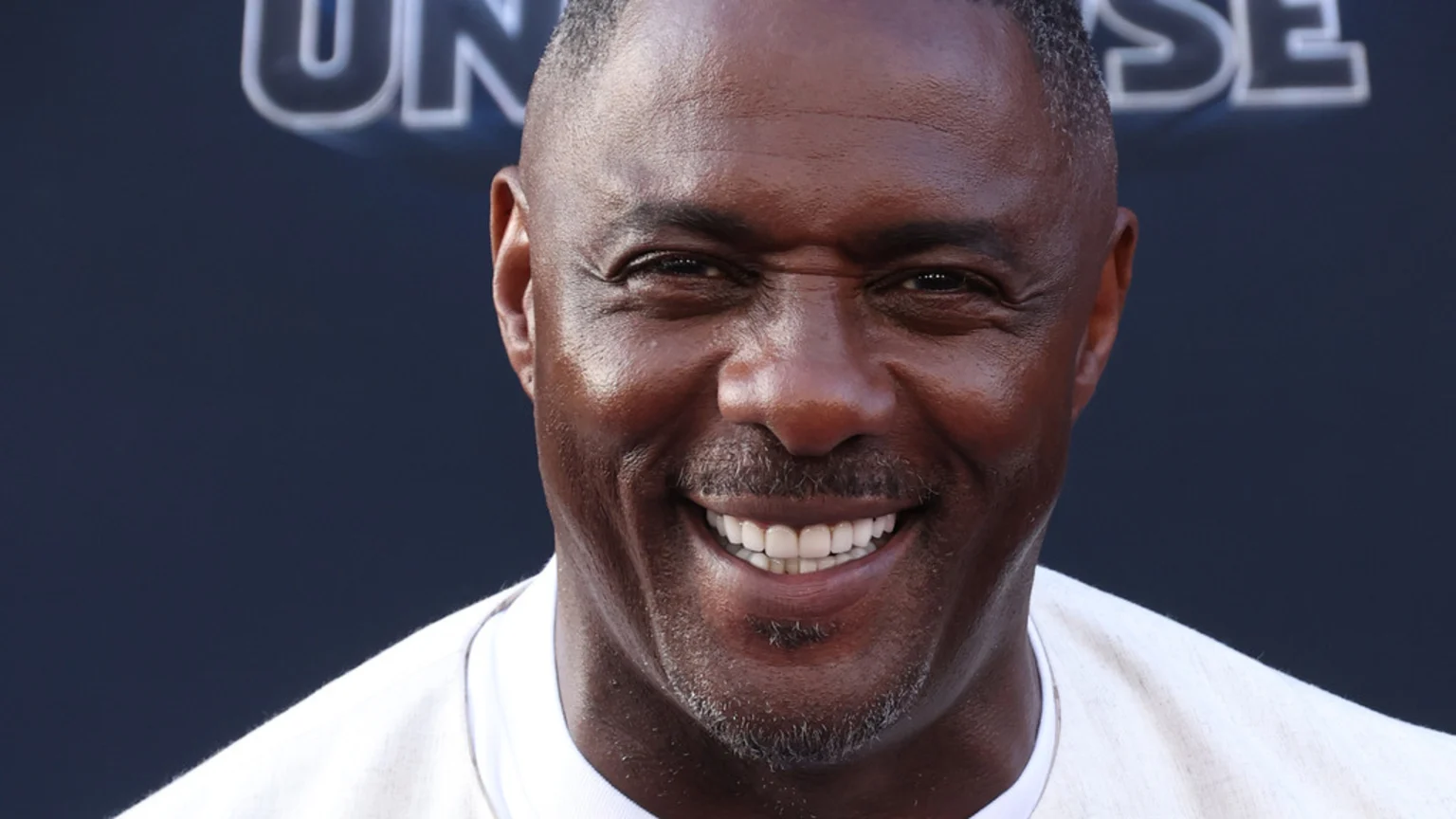 Close-up of actor Idris Elba smiling broadly, with his eyes crinkling, wearing a white shirt against a dark, blurred background with a partial sign visible.