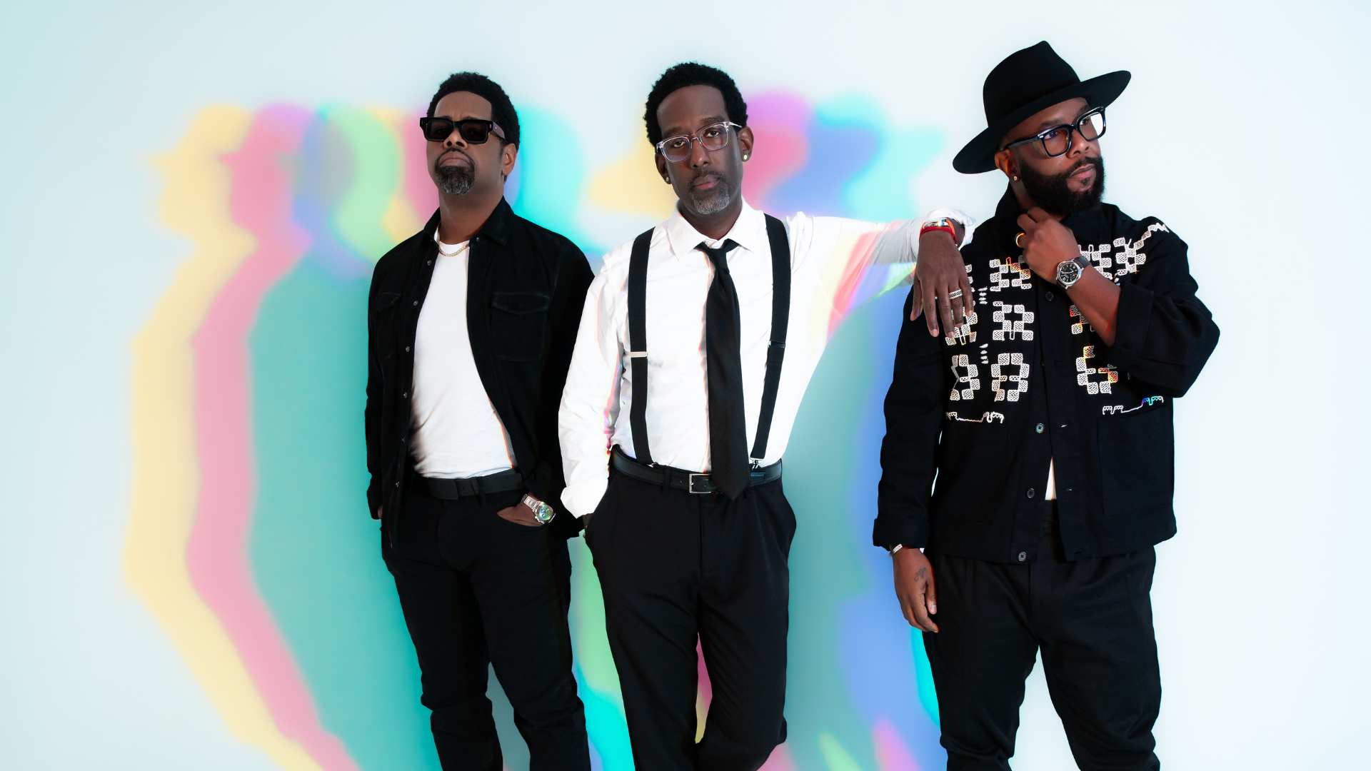 Three stylish Black men pose, one wearing sunglasses, another suspenders, and the third a hat and patterned jacket, against a light blue background with colorful, blurred streaks.