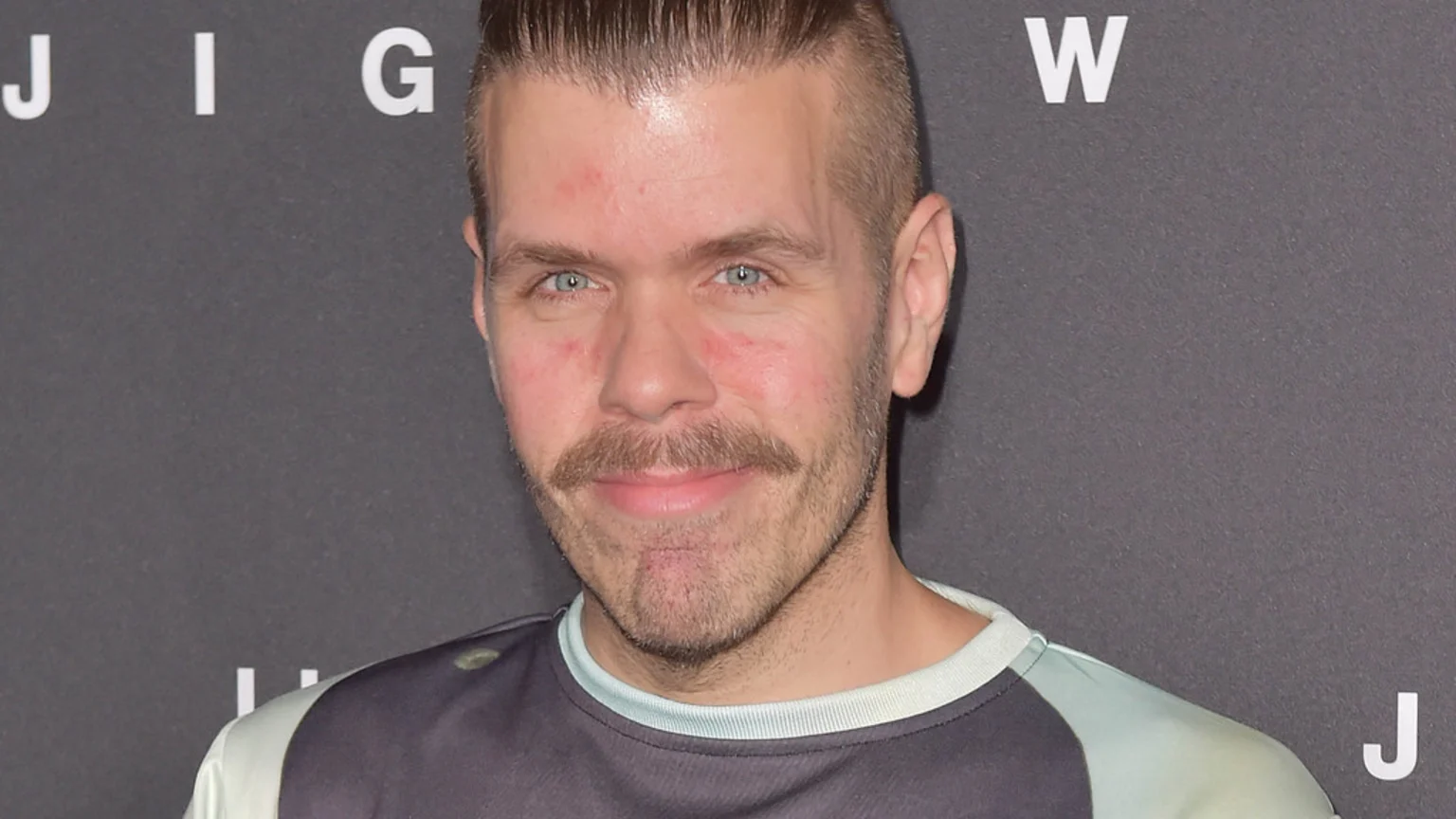 A man with light blue eyes, a handlebar mustache, and slicked-back hair, with some redness on his face, smiles slightly in a two-tone shirt against a dark background with white letters.