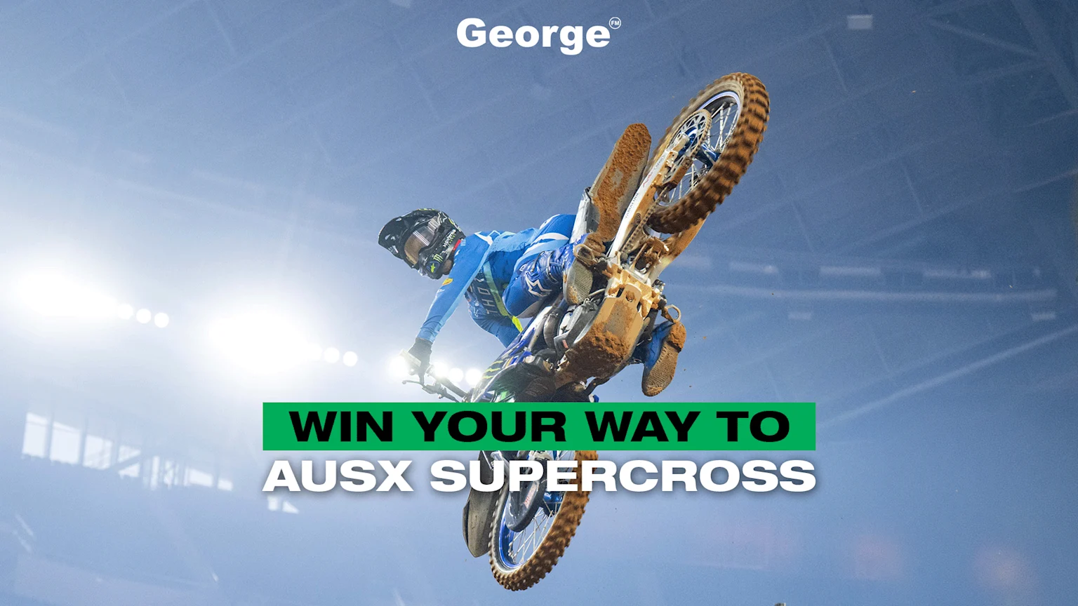 A motocross rider in blue gear performs a jump on a mud-splattered dirt bike in a stadium with bright lights, and promotional text 'WIN YOUR WAY TO AUSX SUPERCROSS' and 'George'.