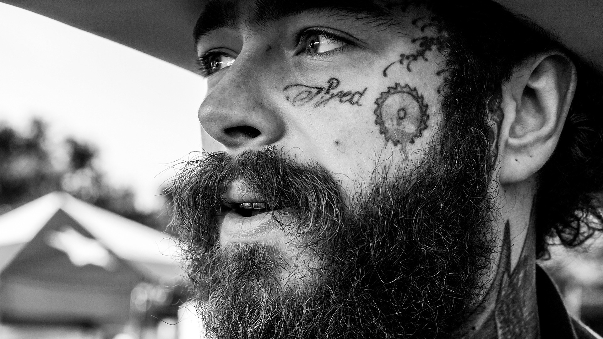 A black and white close-up of Post Malone's face, showing his beard, mustache, face tattoos ("Tired" and a circle with a "P"), and metal teeth as he looks up.