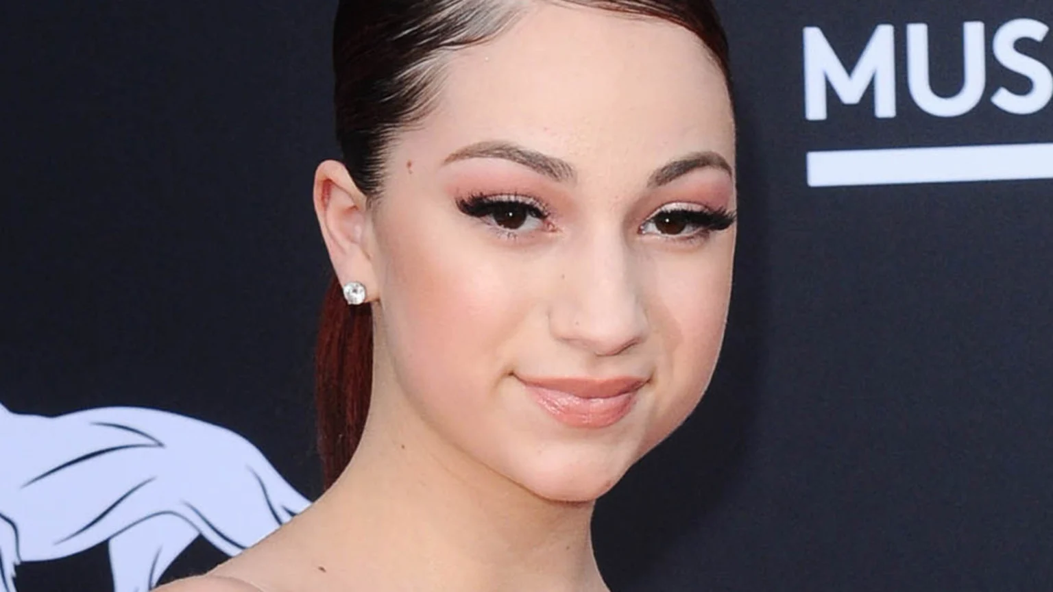 Close-up of Bhad Bhabie, a young woman with dark hair pulled back and styled baby hairs, wearing pink eyeshadow, long dark eyelashes, and glossy nude lips, smiling slightly against a dark background.