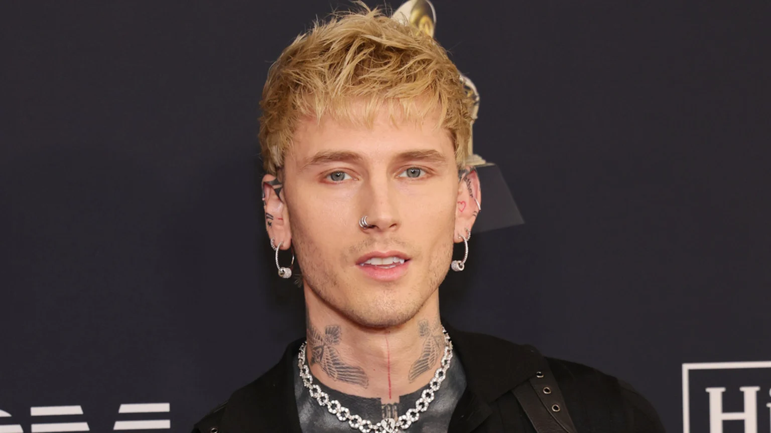 Close-up portrait of Machine Gun Kelly looking at the camera, featuring his blonde hair, light eyes, nose ring, multiple ear piercings, and neck tattoos.