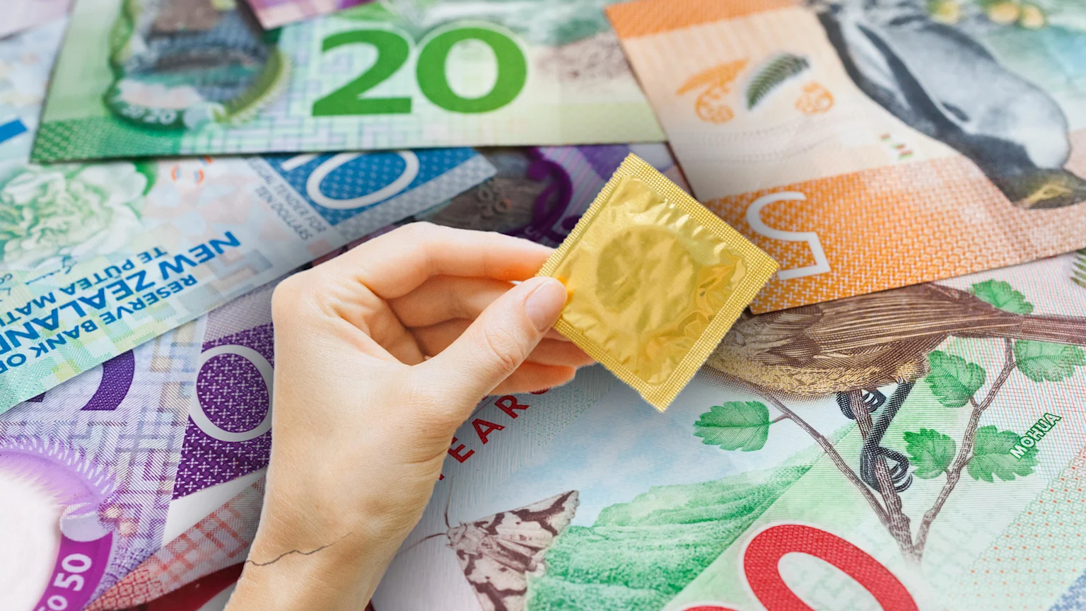 A hand holds a gold-wrapped condom above a collection of scattered New Zealand dollar banknotes, featuring denominations like 5, 10, 20, and 50.