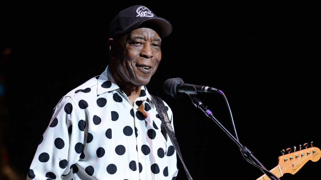 Blues musician Buddy Guy, wearing a white polka dot shirt and black cap, smiles while performing on stage with a microphone and guitar.