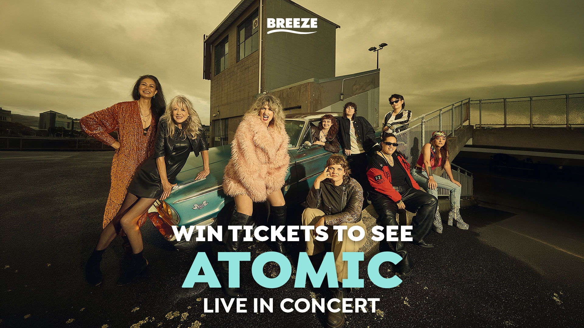 Eight people in stylish outfits pose around a vintage blue car on an urban rooftop under an overcast sky, with the word "ATOMIC" prominently displayed on the image.