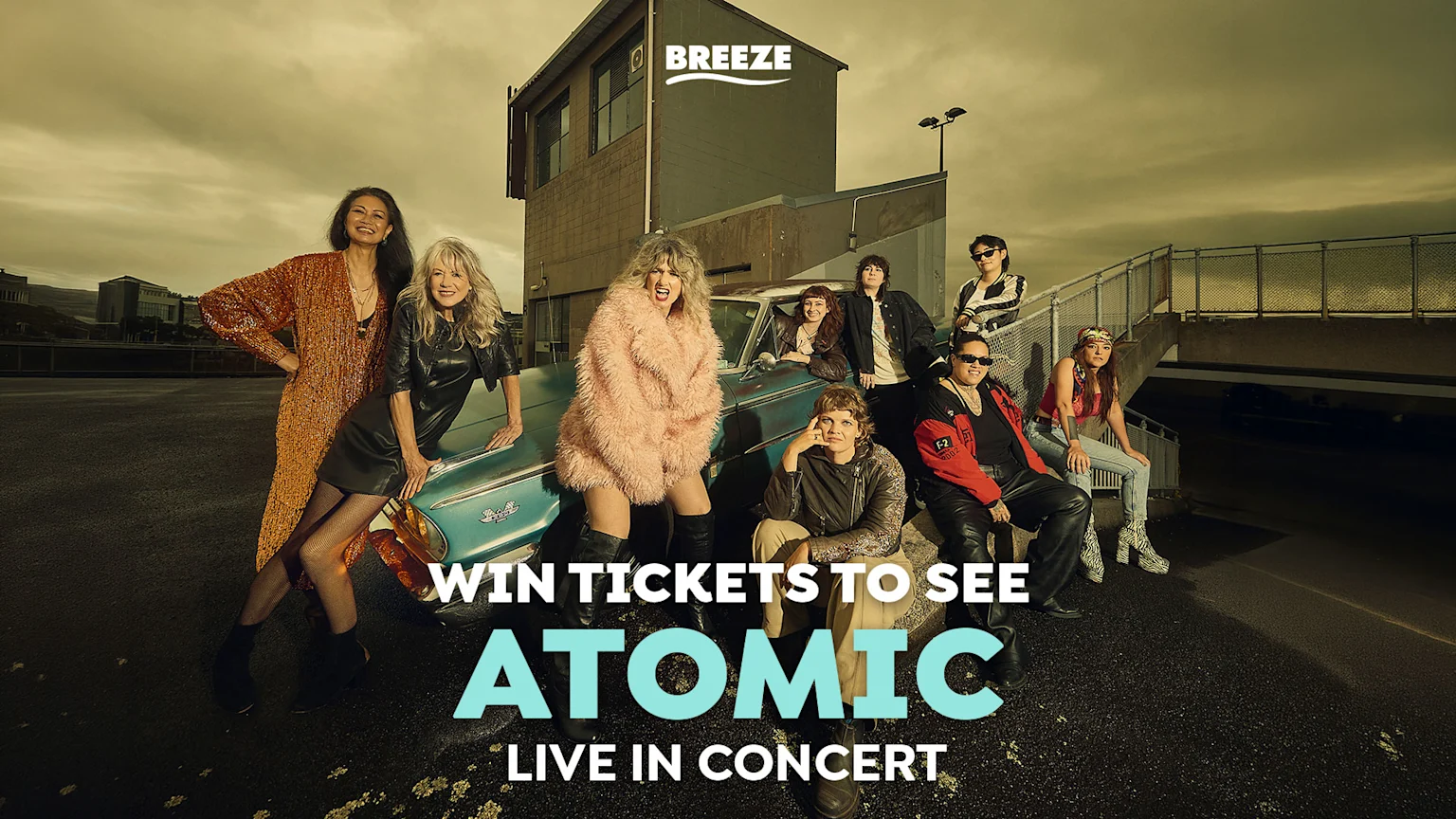 Eight people in stylish outfits pose around a vintage blue car on an urban rooftop under an overcast sky, with the word "ATOMIC" prominently displayed on the image.