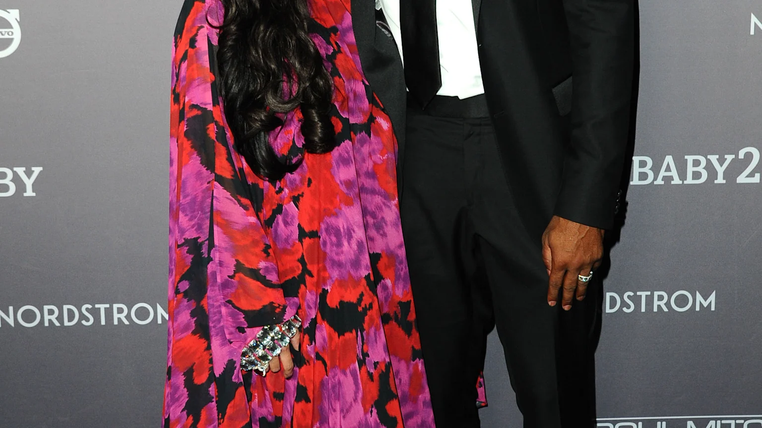 Kobe Bryant, smiling in a black suit, stands next to Vanessa Bryant, who wears a flowing pink, red, and black floral dress with a sparkling necklace.