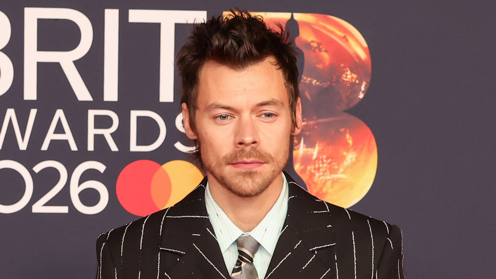 Harry Styles, a man with short brown hair and a light beard, wears a pinstriped suit, light blue shirt, and striped tie, standing in front of a "BRIT AWARDS 2026" backdrop with a Mastercard logo.