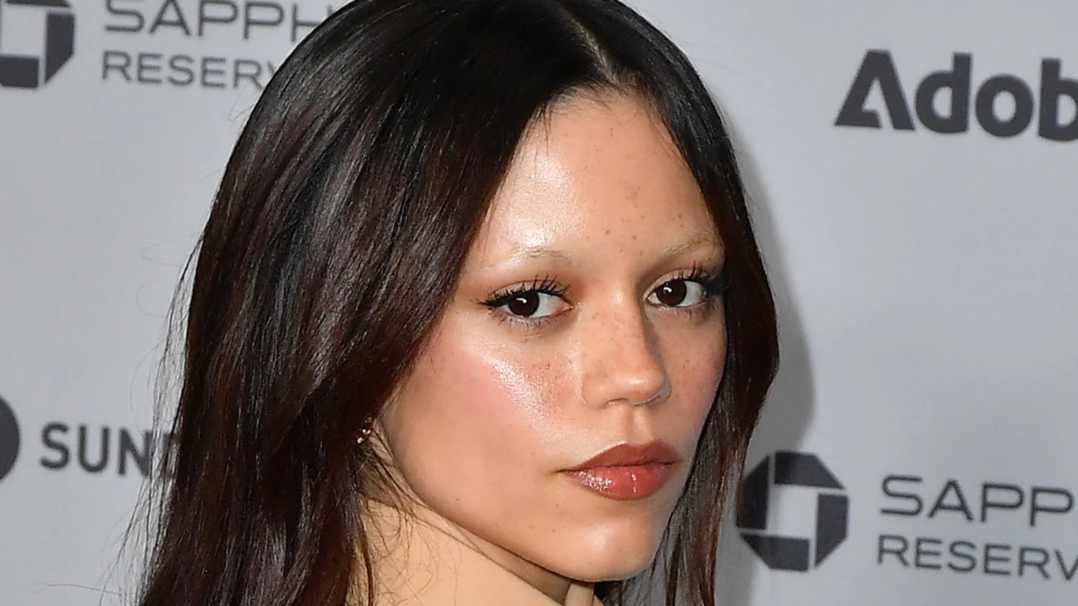 A close-up of Jenna Ortega shows her with dark hair, light eyebrows, freckles, and glossy lips, looking slightly to the side against a blurred white background.