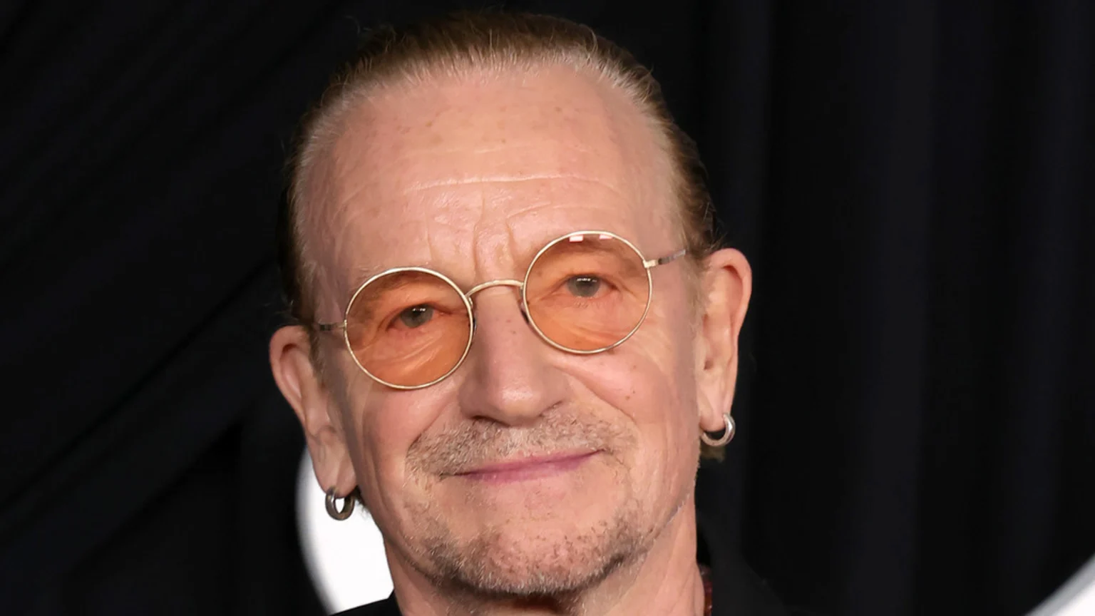 Close-up of a man with a goatee, amber-tinted round glasses, and silver hoop earrings, looking directly at the camera with a subtle smile against a dark background.