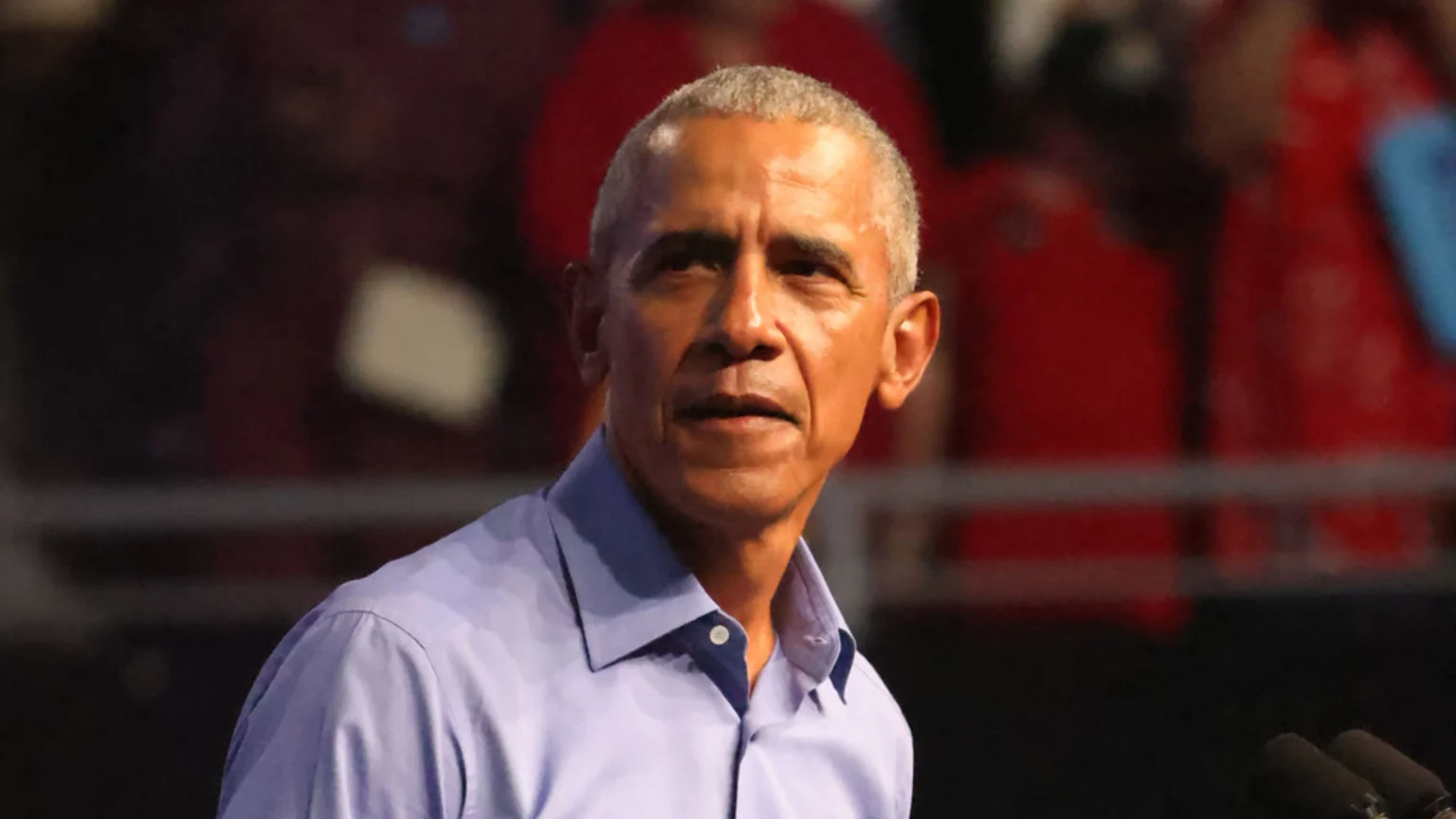 A medium close-up image of Barack Obama at an indoor event venue, wearing a light blue collared shirt and displaying a focused expression. Two dark microphones are visible at the bottom right corner against a blurred dark background.