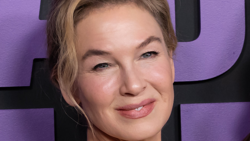 A close-up of Renee Zellweger smiling slightly, with blue eyes and blonde hair, against a purple and black background with partial letters visible.