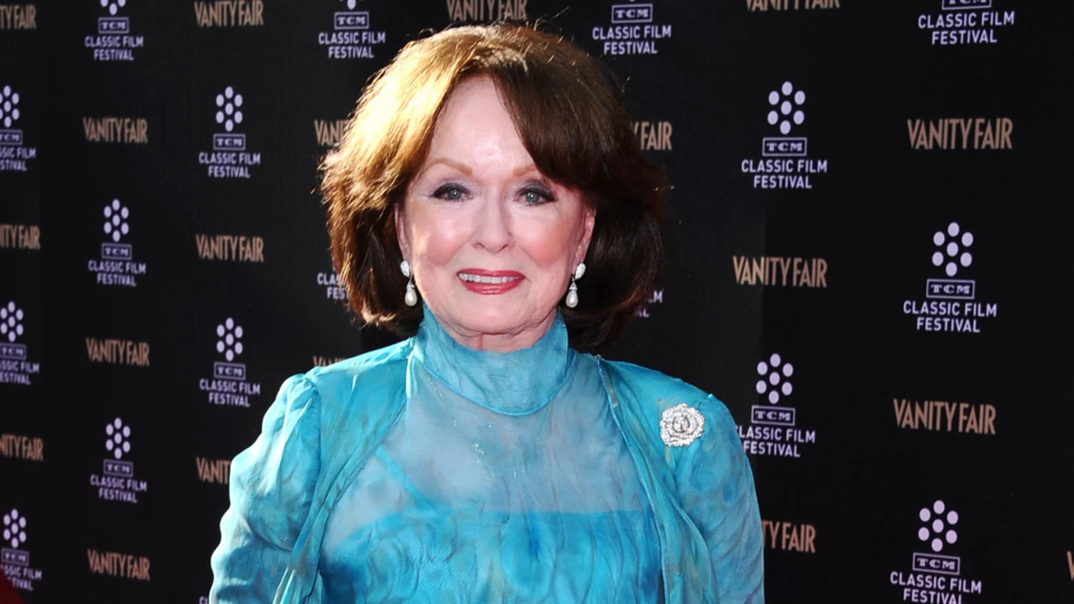Actress Ann Blyth smiles, wearing a teal high-neck blouse, a brooch, and pearl earrings, in front of a black backdrop featuring repeated Vanity Fair and TCM Classic Film Festival logos.