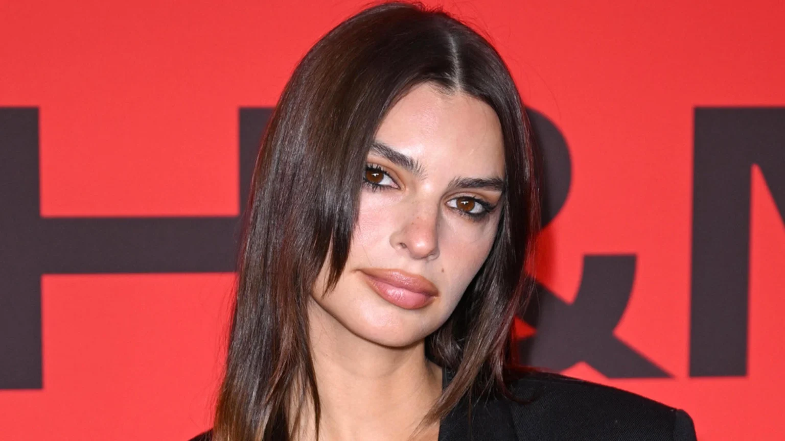 Close-up portrait of Emily Ratajkowski with long dark hair, brown eyes, and neutral lipstick, wearing a black blazer, against a red background with blurry dark letters.