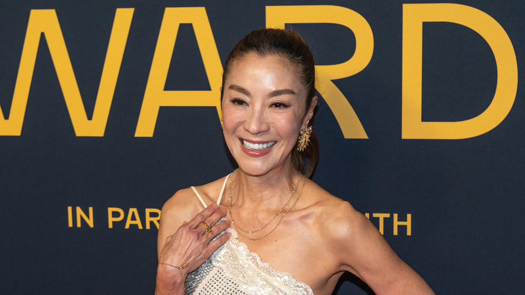 Michelle Yeoh smiles warmly, facing slightly left, in front of a dark blue background with yellow letters spelling "WARD" and "IN PARIS WITH", wearing a white, sparkly, one-shoulder top, layered necklaces, and a large gold earring.